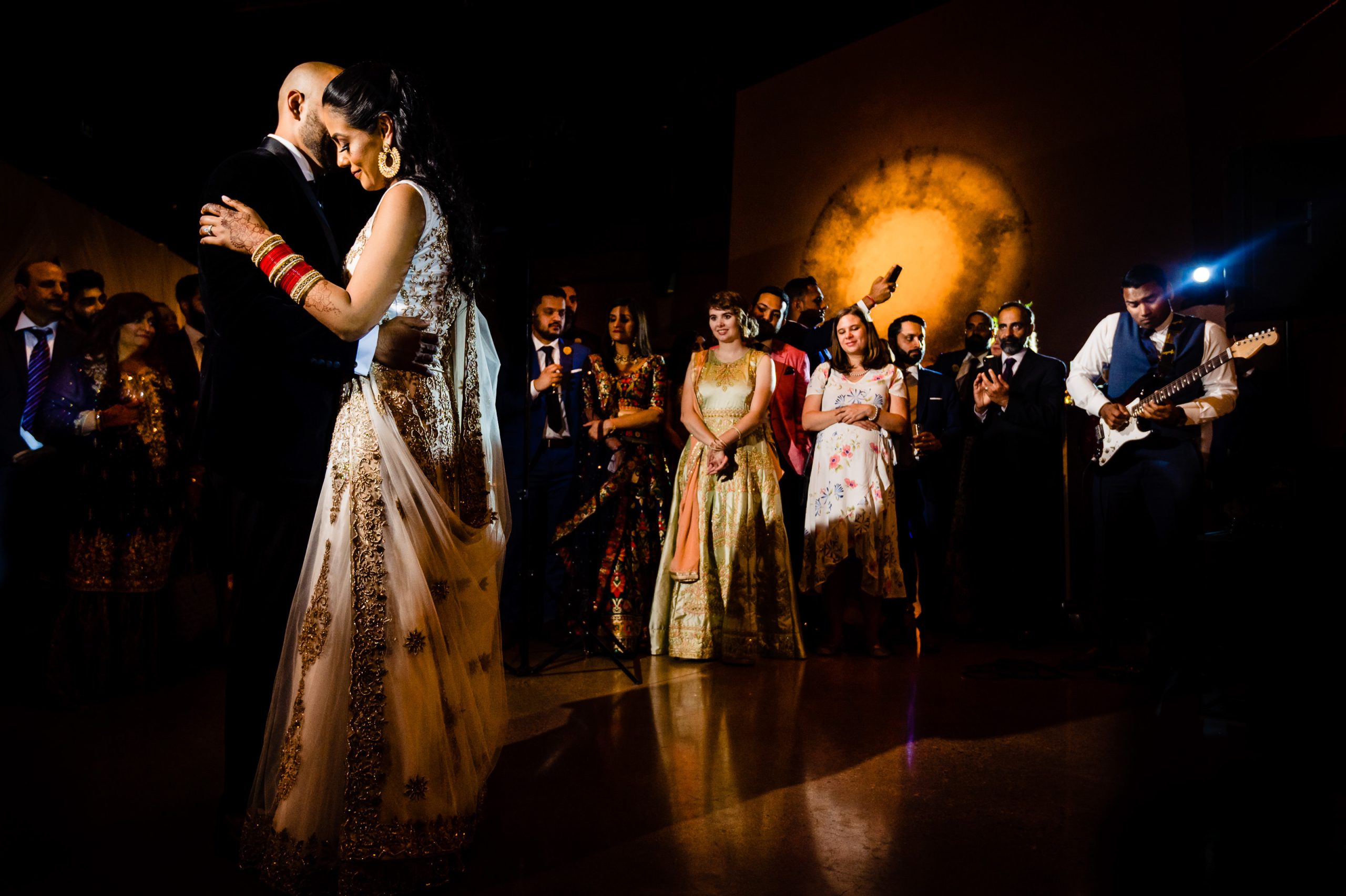 A couple shares a tender dance at a wedding reception, surrounded by elegantly dressed guests. The bridal party stands in the background, watching with joyful expressions. The dim lighting casts a romantic glow, highlighting the couple in the center.