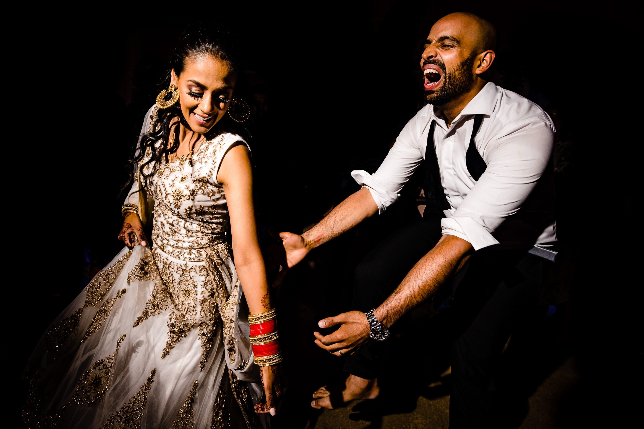 A joyful couple dances energetically at a celebration. The woman, wearing a traditional embroidered dress and gold jewelry, laughs with joy. The man, dressed in a white shirt and black suspenders, sings and extends his arms in a lively gesture.