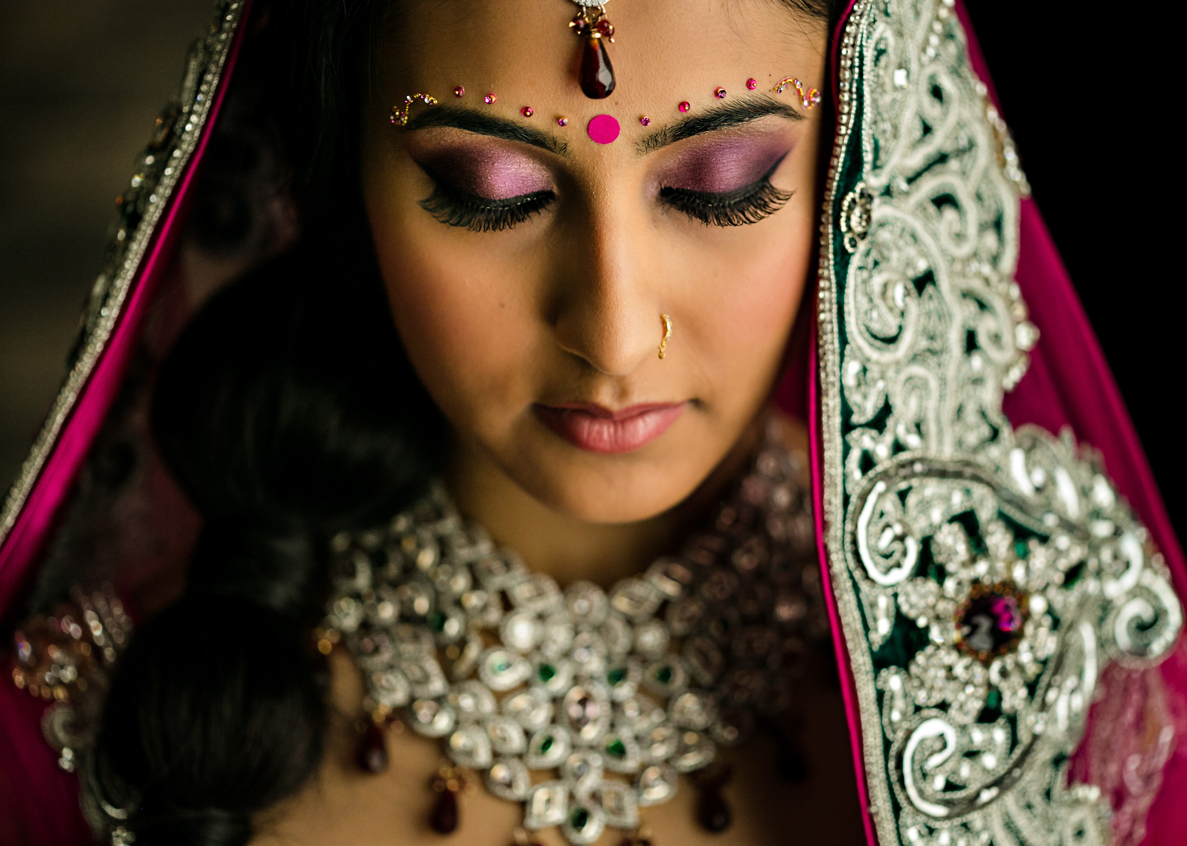 Close-up of a bride with closed eyes, adorned in traditional bridal attire. She wears a jeweled headpiece, colorful eye makeup, intricate jewelry, and a detailed embroidered dupatta draped over her head. The image captures her serene and graceful demeanor.