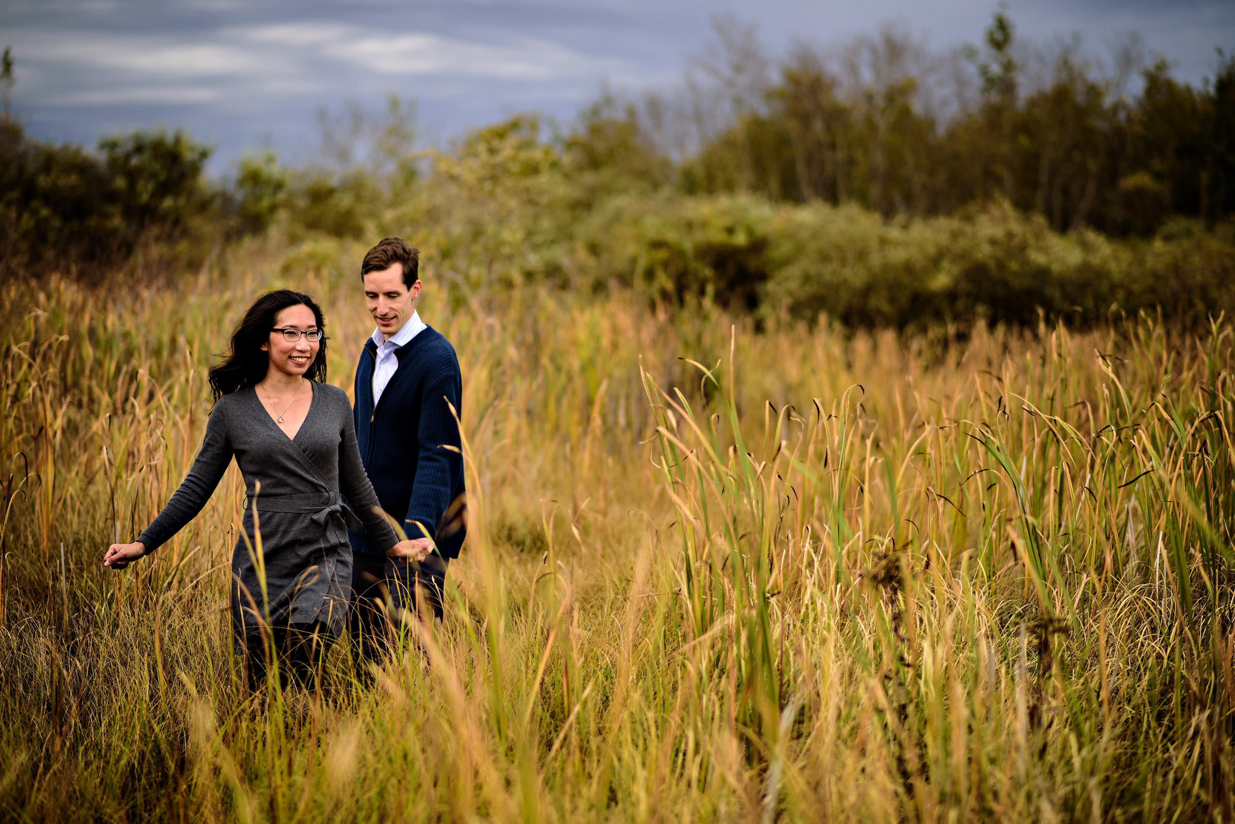 A couple strolls through a tall grassy field under Winnipeg's cloudy wedding sky.