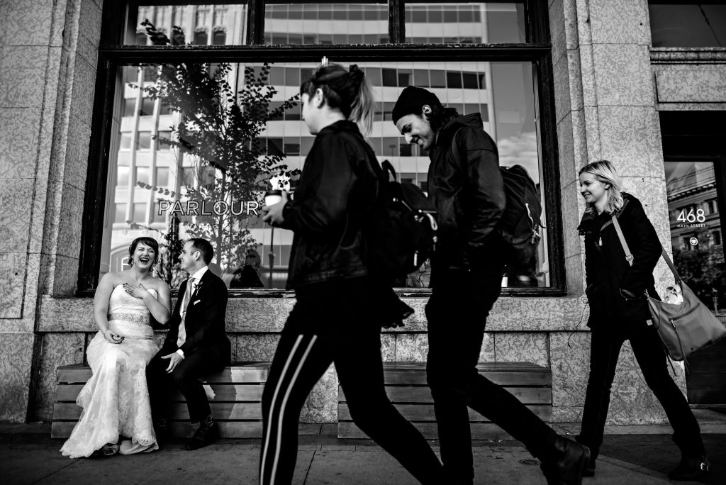Ab, bride with her groom, laughing on a bench on a sidewalk in downtown Winnipeg, while people walk in front of them