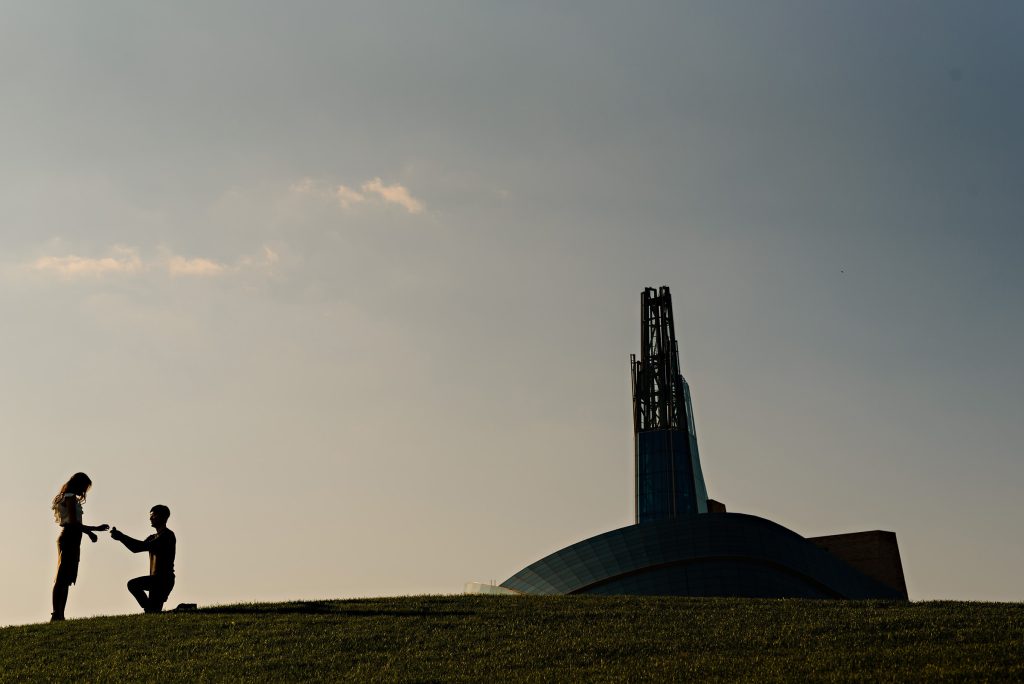 Silhouette photo of a man on his knees, proposing to his fiancé with the Canadian museum of human rights in the background at the forks