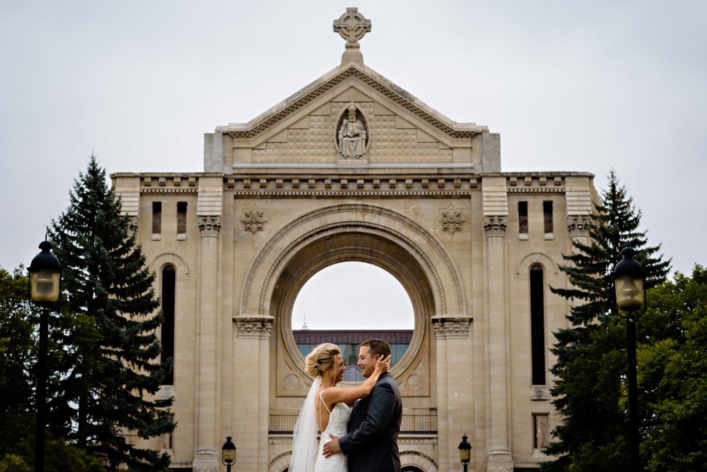 A bride and groom, embracing and smiling at each other in front of Saint Boniface Cathedral in Winnipeg, Manitoba