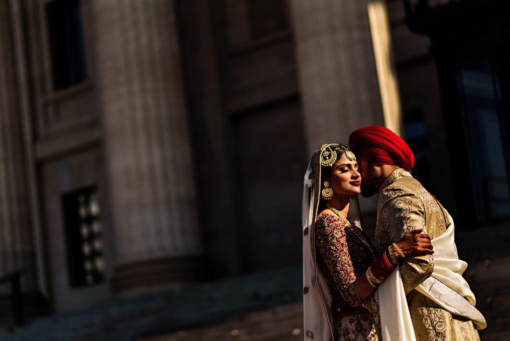 An Indian bride and groom, embracing for a wedding photograph in front of the Winnipeg legislative building in the beautiful sunlight
