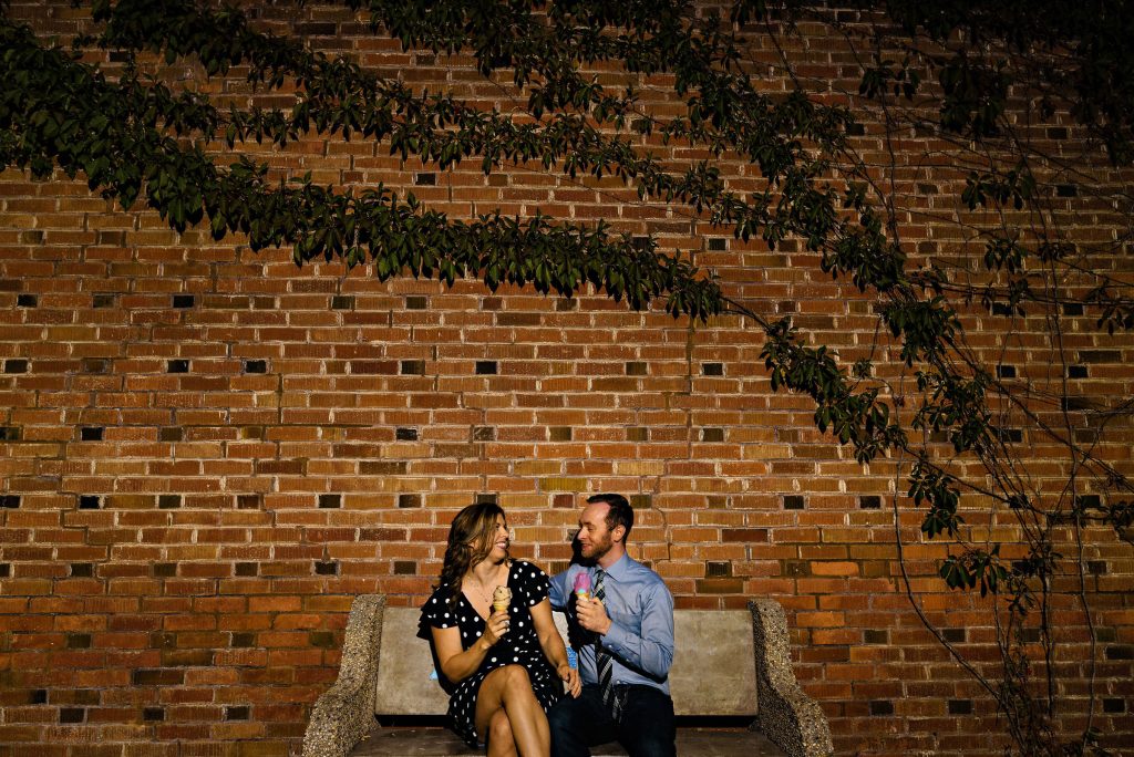 an Engaged couple sitting on a bench eating ice cream in front of a brick wall in Winnipeg Manitoba