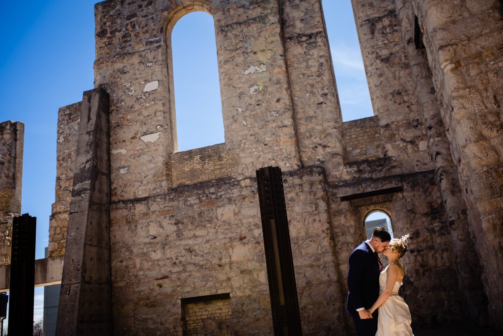 Bride and groom, embracing and kissing in front of a stone wall at Saint Boniface Cathedral in Winnipeg