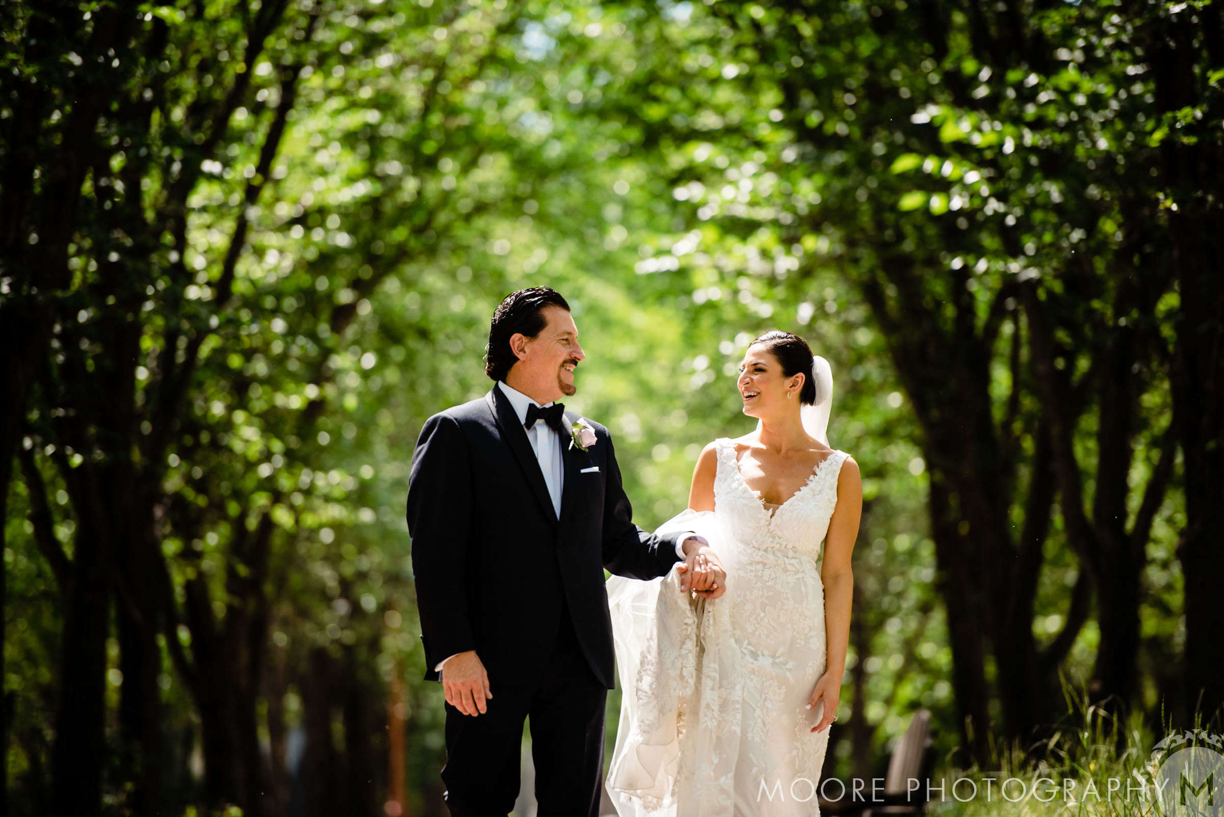 A bride and groom holding hands on their wedding day, laughing, and smiling with trees in the background at the forks