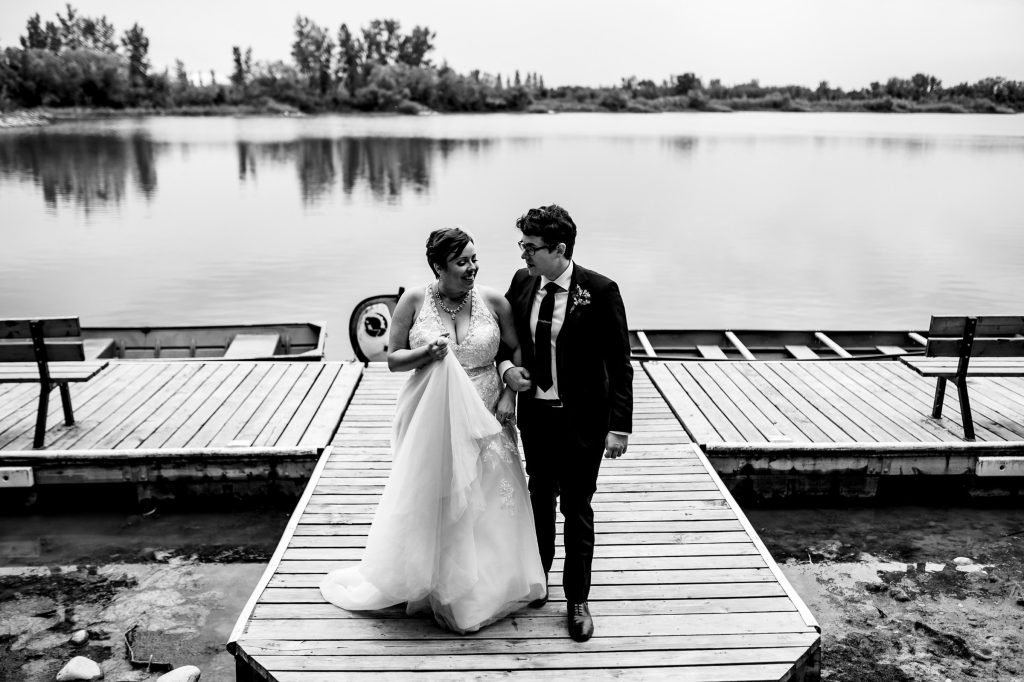 Bride and groom, walking and talking on a dock, with a lake in the background in Winnipeg Manitoba