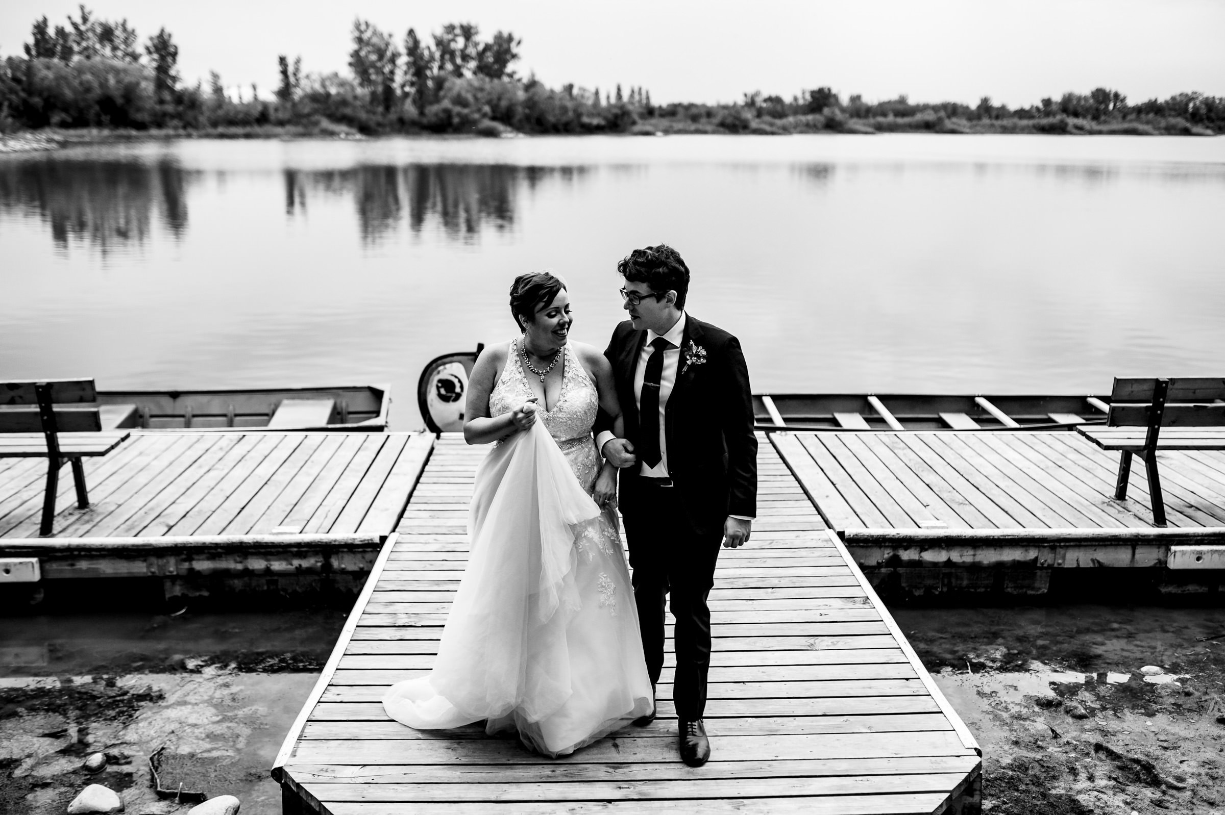 Bride and groom stroll hand in hand on a scenic Winnipeg lakeside dock.