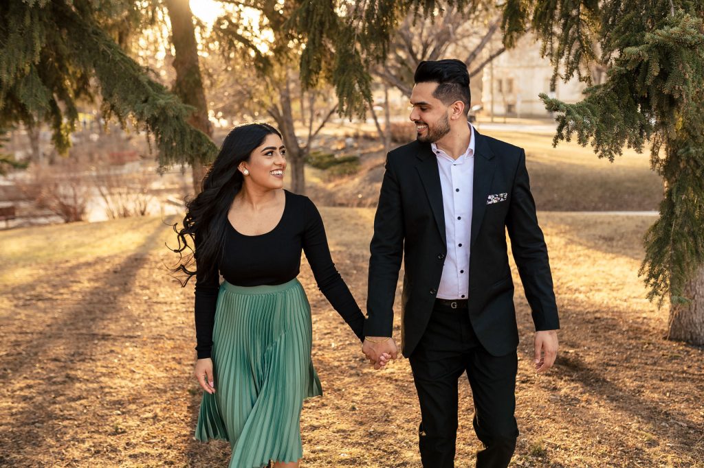 I engaged couple walking and holding hands while smiling at each other through a forest in Winnipeg