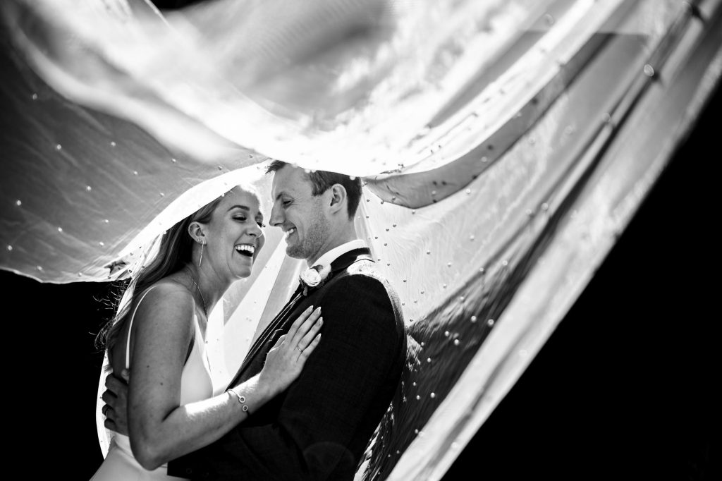 Bride and Groom laughing under the brides wedding vale in the sunlight