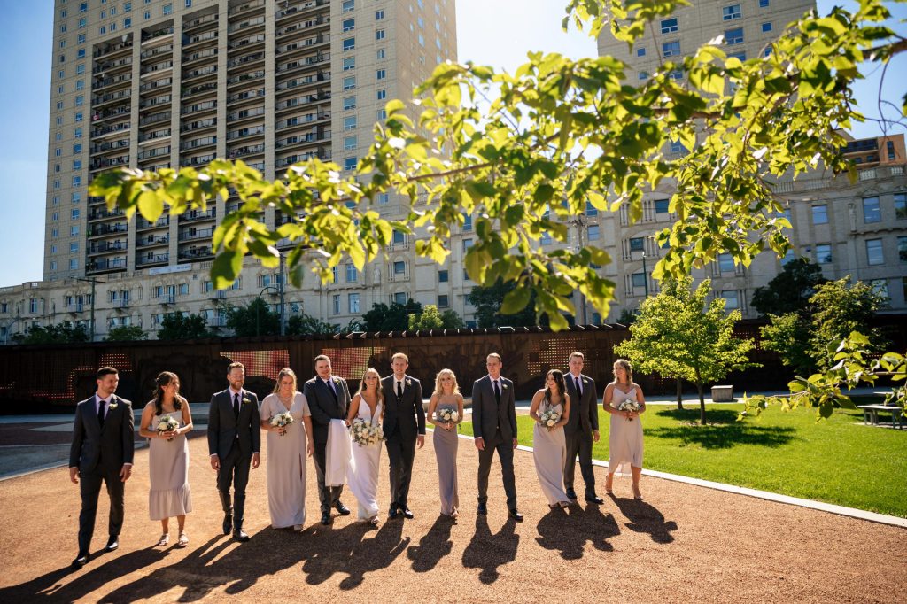 A bride and groom with their wedding party, walking in the sunlight under tree branches at upper Fort Gary in Winnipeg Manitoba