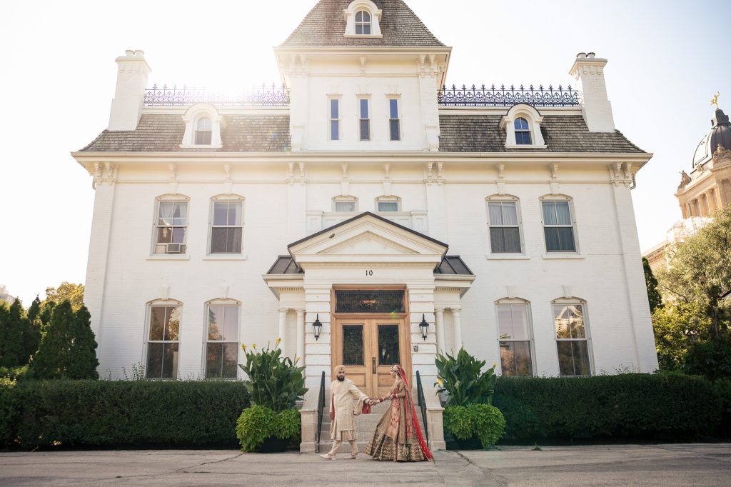 An Indian bride and groom walking on their wedding day with a big white house in the background