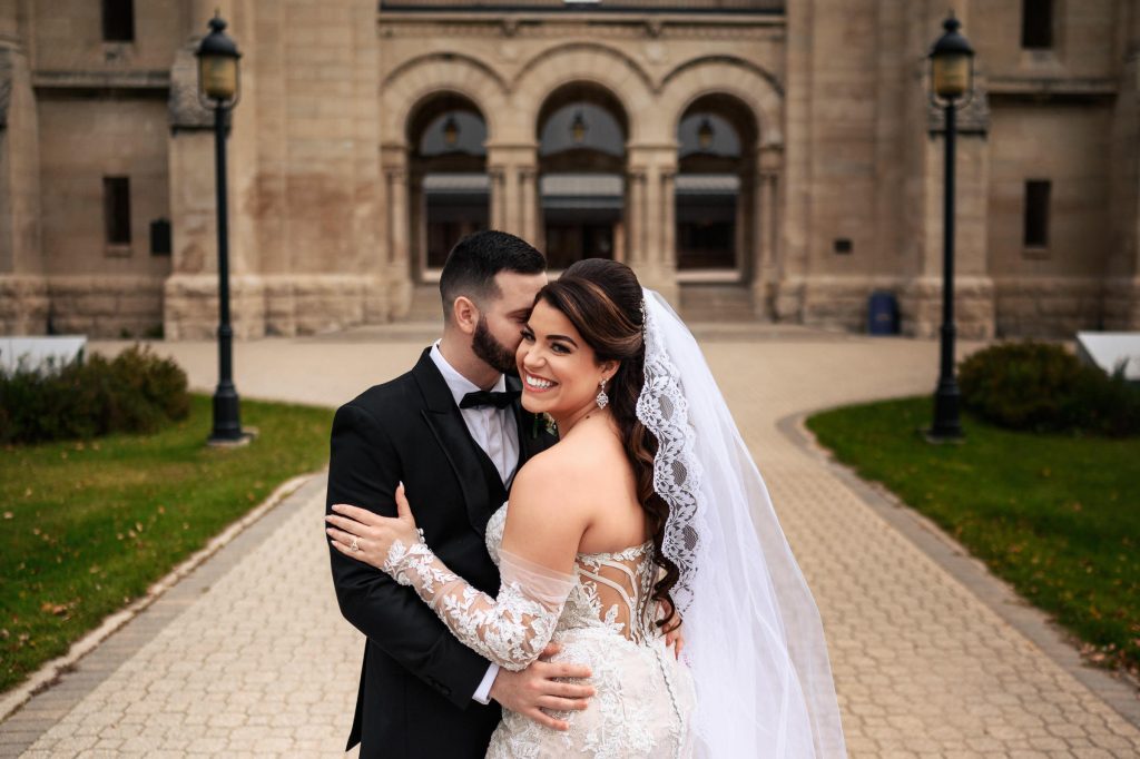 Bride and groom, embracing and posing for a photo in front of Saint Boniface Cathedral in Winnipeg