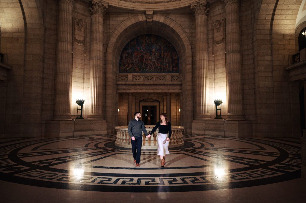And engaged, holding hands walking through the Winnipeg legislative building in beautiful architecture