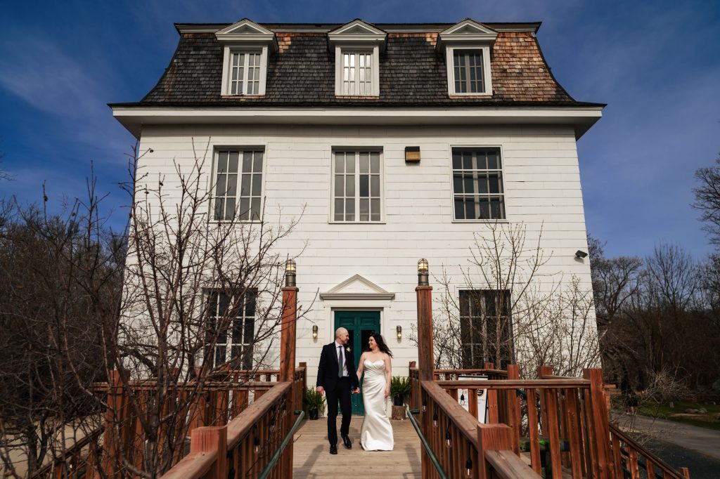 A bride and groom holding hands and walking away from a white house under a blue sky