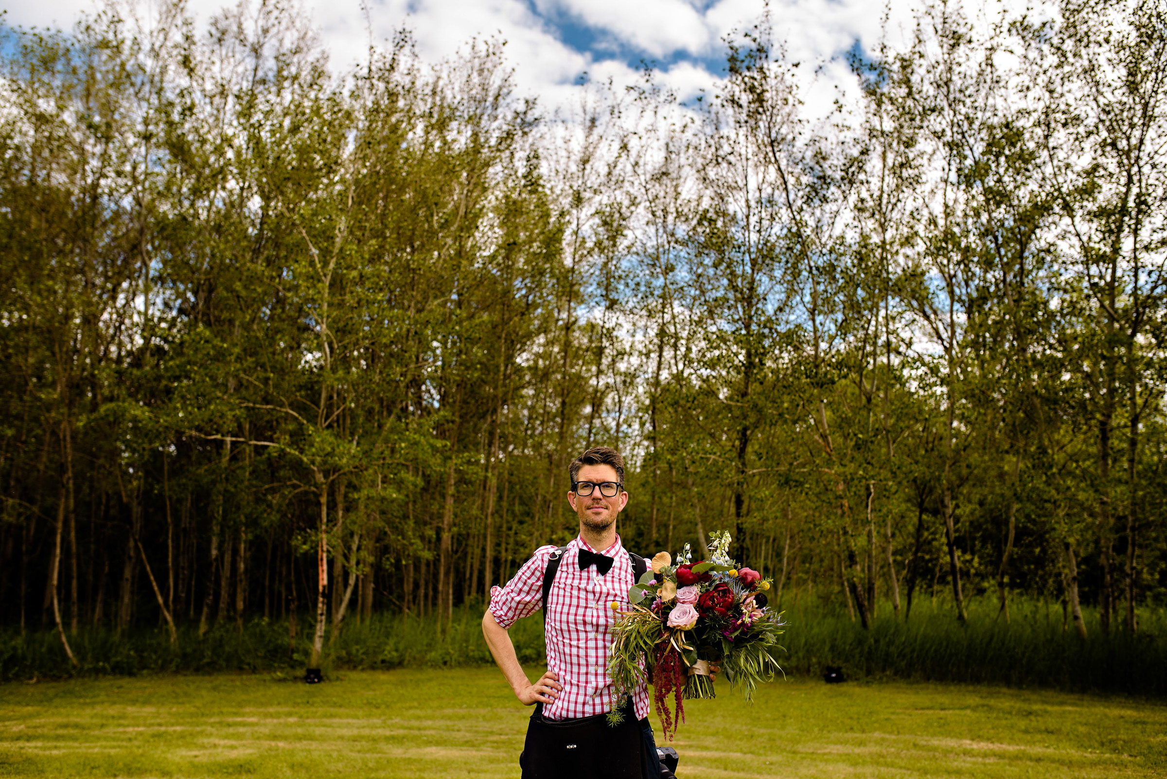 A person with glasses stands confidently in a field, wearing a pink checkered shirt and bow tie, holding a large, colorful bouquet of flowers. The background features a line of trees and a partly cloudy sky.