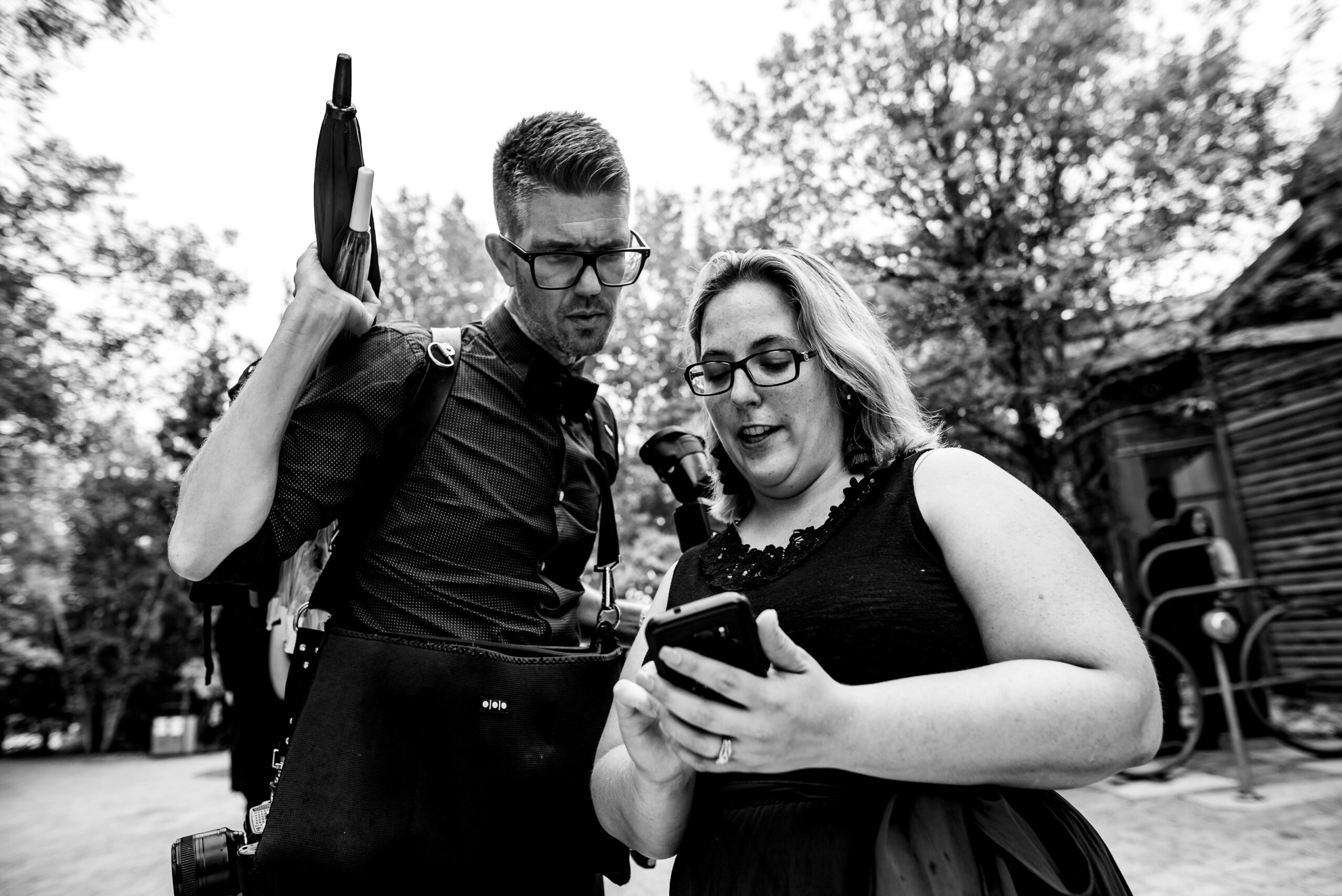 A black-and-white photo of two people outdoors. They are both looking at a smartphone the woman is holding. The man, wearing glasses and carrying a bag and umbrella, looks over her shoulder. The background has trees and possibly some people in the distance.