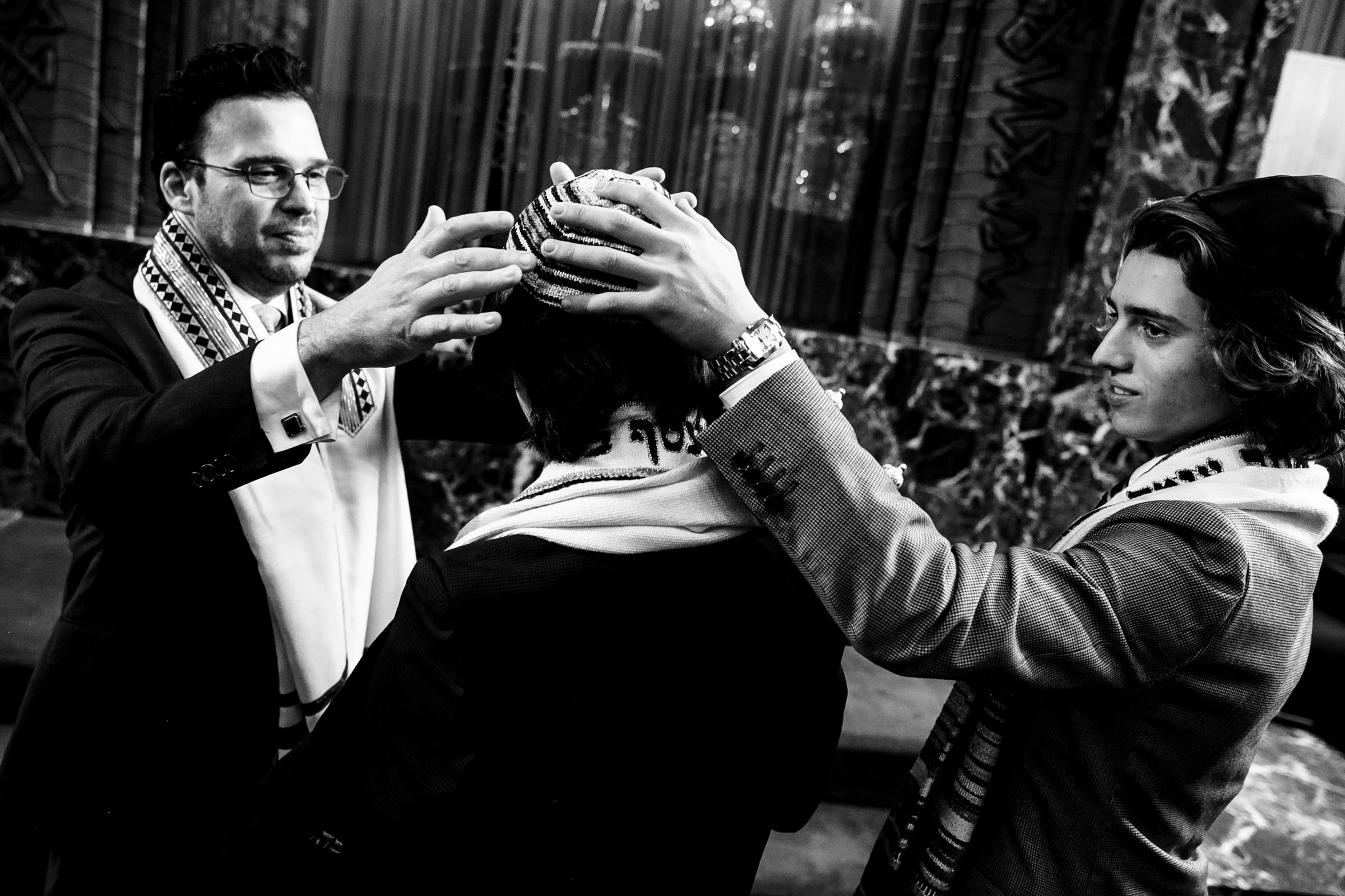 Three men are participating in a religious ceremony. The man on the left is raising his hands over the head of the central figure, who is wearing a kippah. The man on the right is also participating, helping to adjust the kippah on the central figure's head.