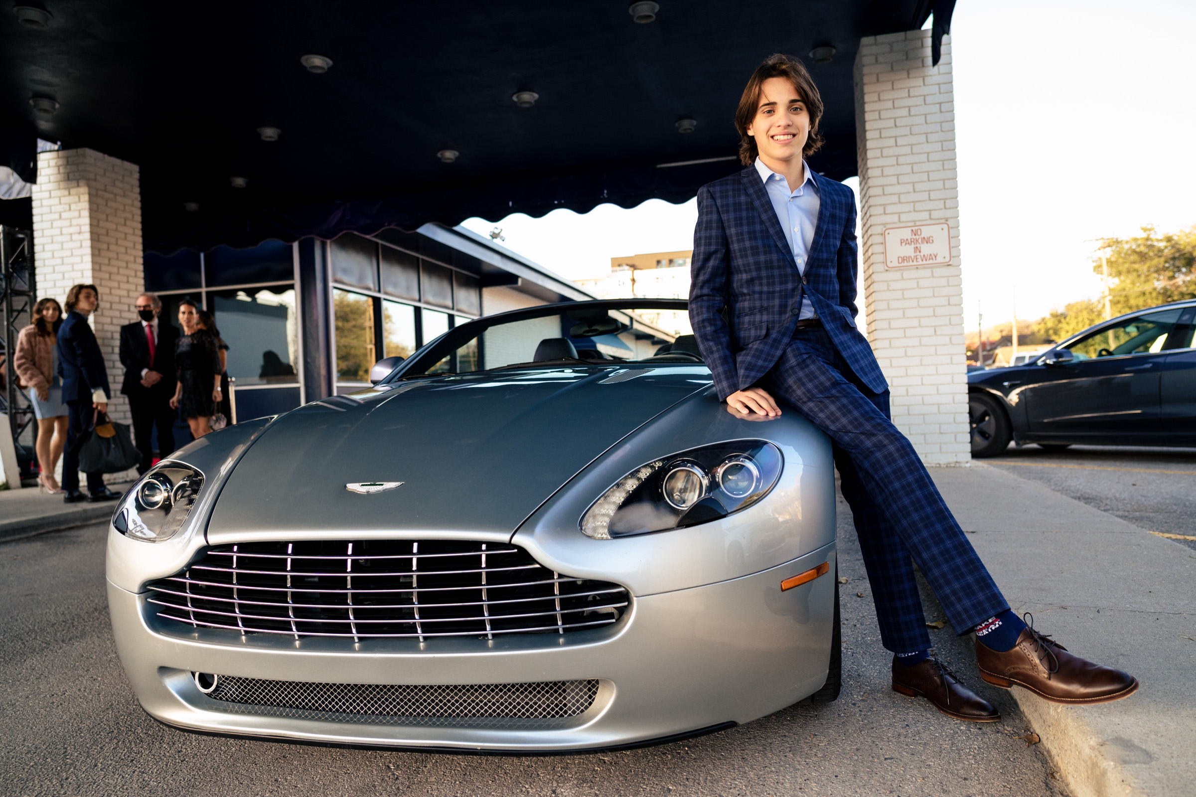 A person in a blue plaid suit is sitting on the hood of a silver Aston Martin sports car. They are smiling and looking at the camera. A few people are gathered in the background near a building entrance. A "One Way Do Not Enter" sign is visible on a nearby pillar.