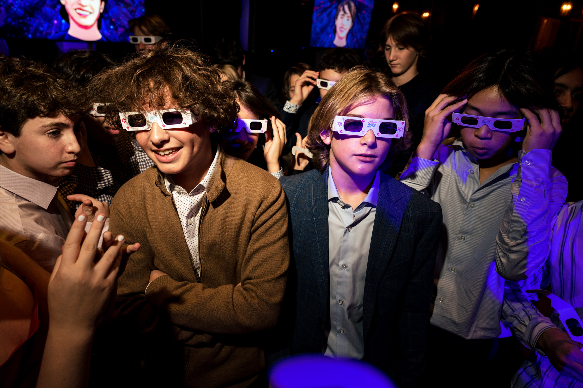 A group of young people stand together, wearing white 3D glasses. The scene is lit with purple and blue lights, creating a festive atmosphere. One person looks at their phone while others adjust their glasses, and a background screen displays a smiling face.