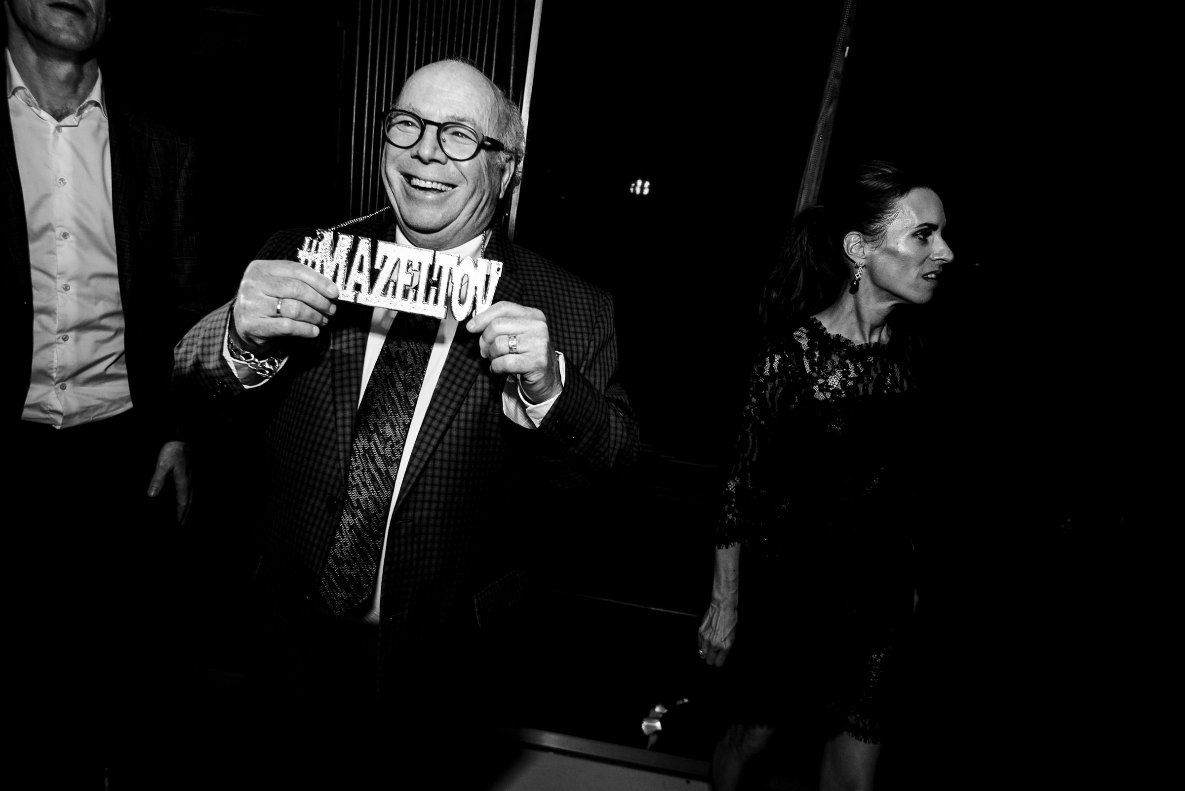 A man wearing glasses and a suit smiles while holding up a sign that reads "#MAZALTOY." A woman in a dark dress stands nearby. The scene is set indoors at an event or celebration with a dark background.