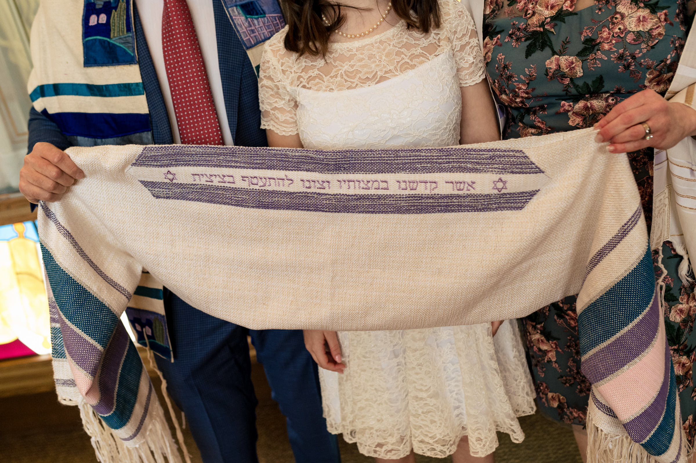 Three people, partially visible, are holding a white prayer shawl with purple and blue accents and Hebrew text. One person is in the center wearing a white lace dress, while the others wear suits and floral print dresses. A colorful, stained glass window is seen in the background.