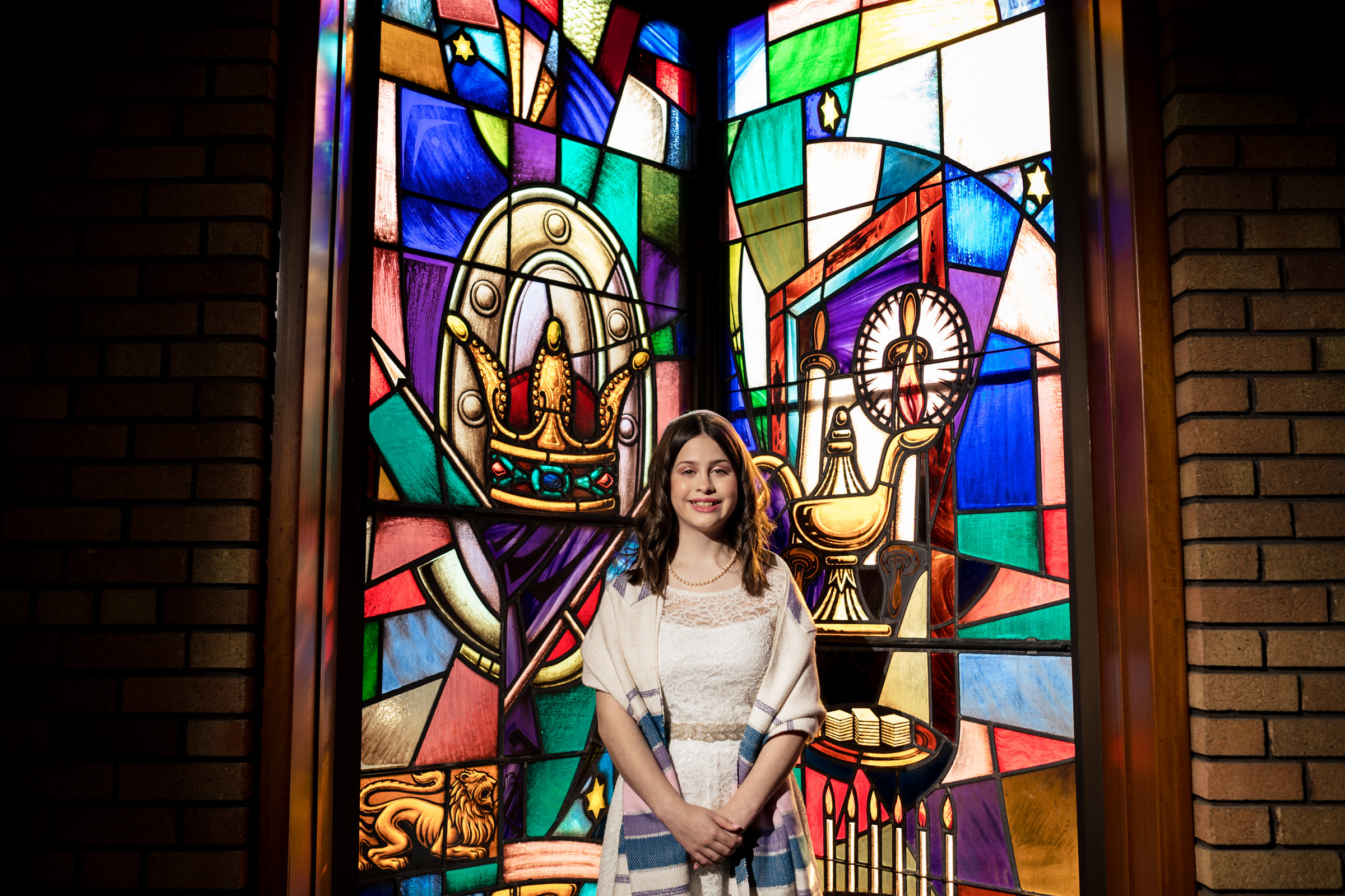A young woman stands in front of a vibrant stained glass window displaying various colorful designs, including a crown and a chalice. She is dressed in a white dress with a blue-striped shawl, smiling, with her hands clasped in front of her.