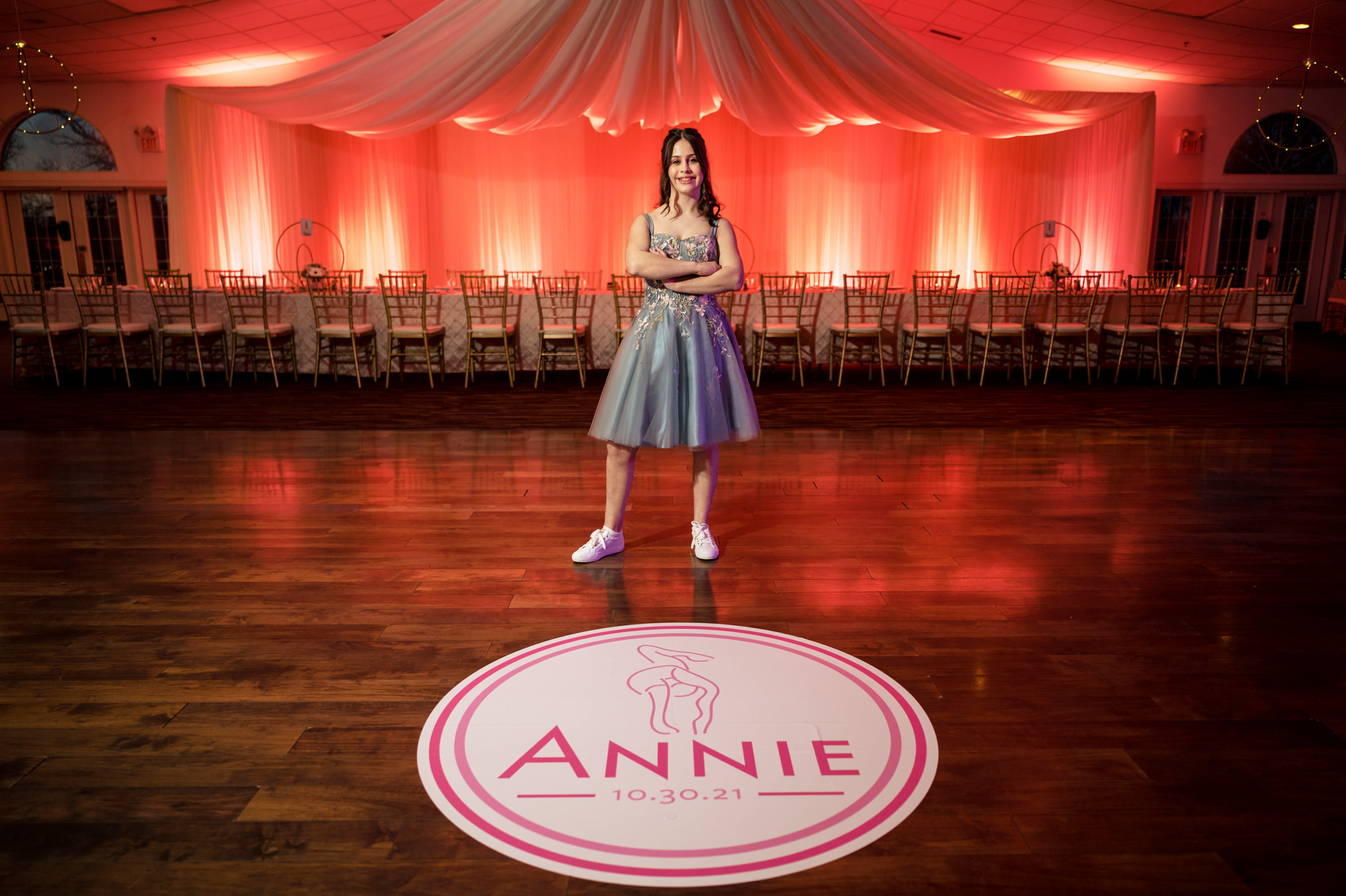 A girl in a silver dress and white sneakers stands confidently on a dance floor with a custom circular mat that reads "Annie 10.30.21." The background features a long dining table, chairs, and elegant drapery with pink lighting.