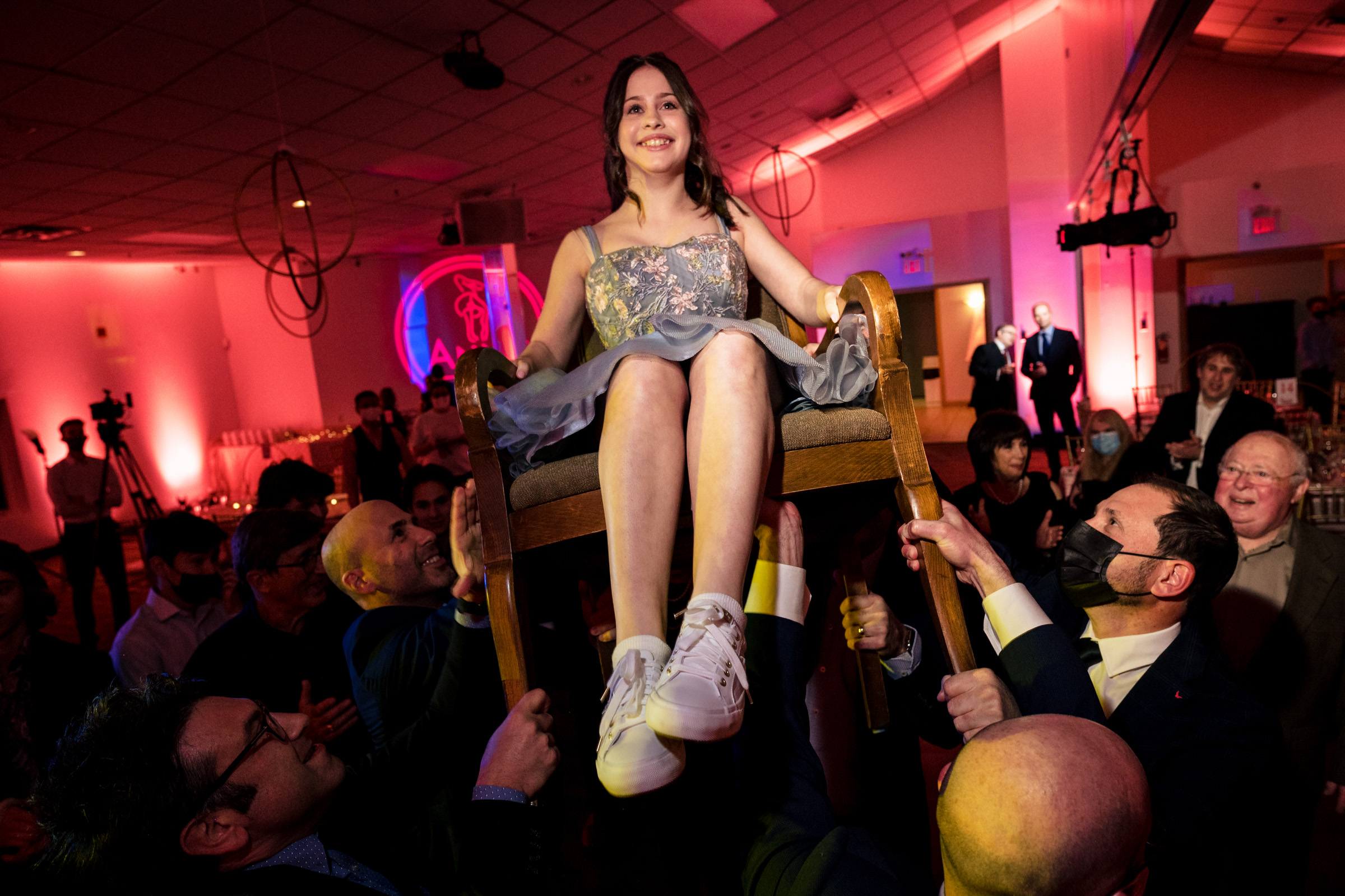 A young girl in a dress and sneakers is joyfully lifted in a chair by several people during a celebration inside a vibrant, warmly lit venue. Onlookers smile and take photos, adding to the festive atmosphere.