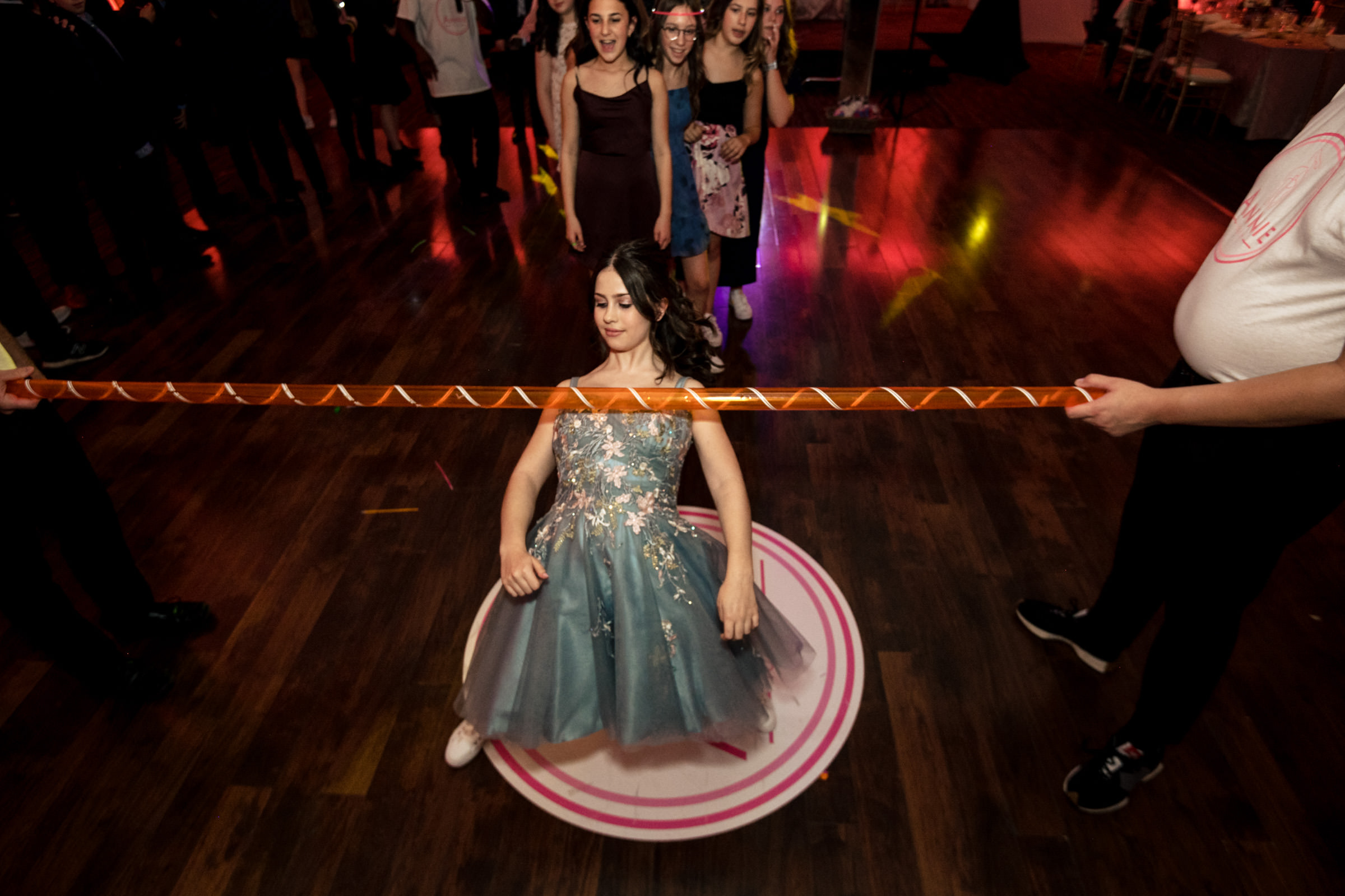 A young woman in a blue dress performs a limbo dance at an event, bending backward low under a limbo stick held by two people. Other attendees watch and cheer in the background as colorful lights illuminate the room.