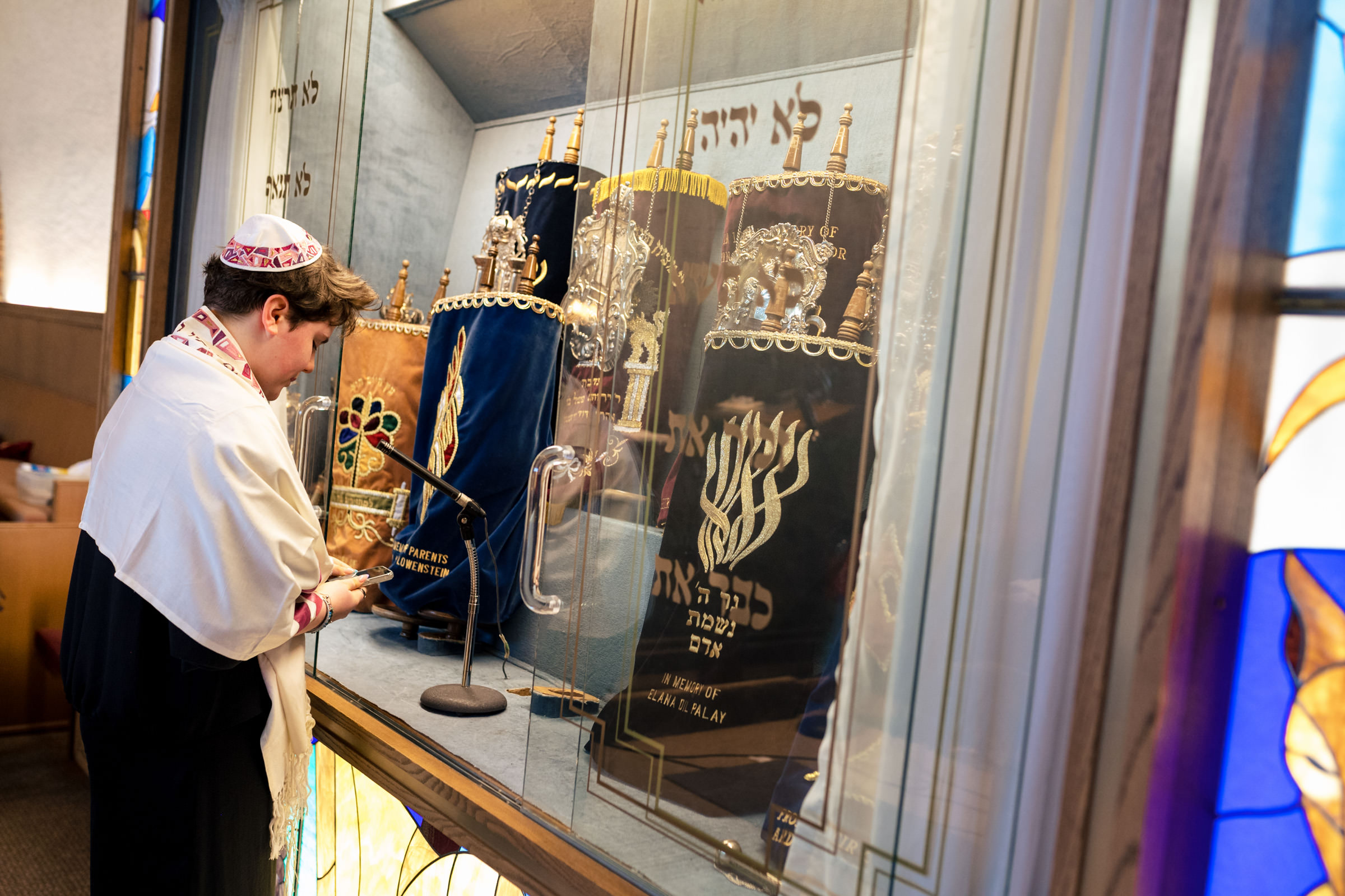 A person wearing traditional Jewish attire stands in front of an ark containing Torah scrolls inside a synagogue. The ark doors are open, revealing ornately adorned Torah scrolls. The individual appears engaged in a religious ritual or prayer.