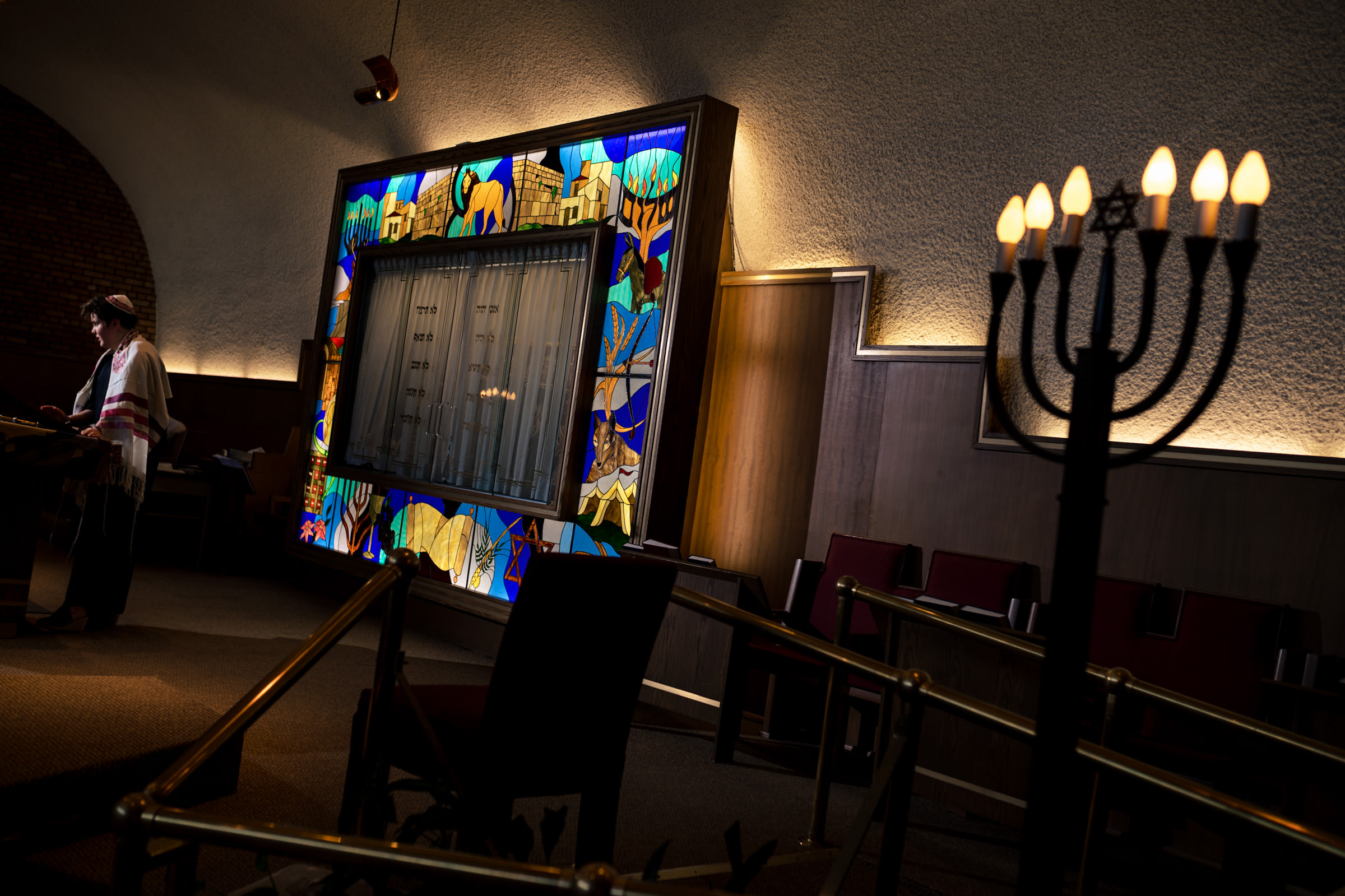 A person reads from a book in a dimly lit room with a large, colorful stained glass window. The window features various abstract designs. In the foreground, there's a menorah on the right side, and several chairs are arranged around the room.