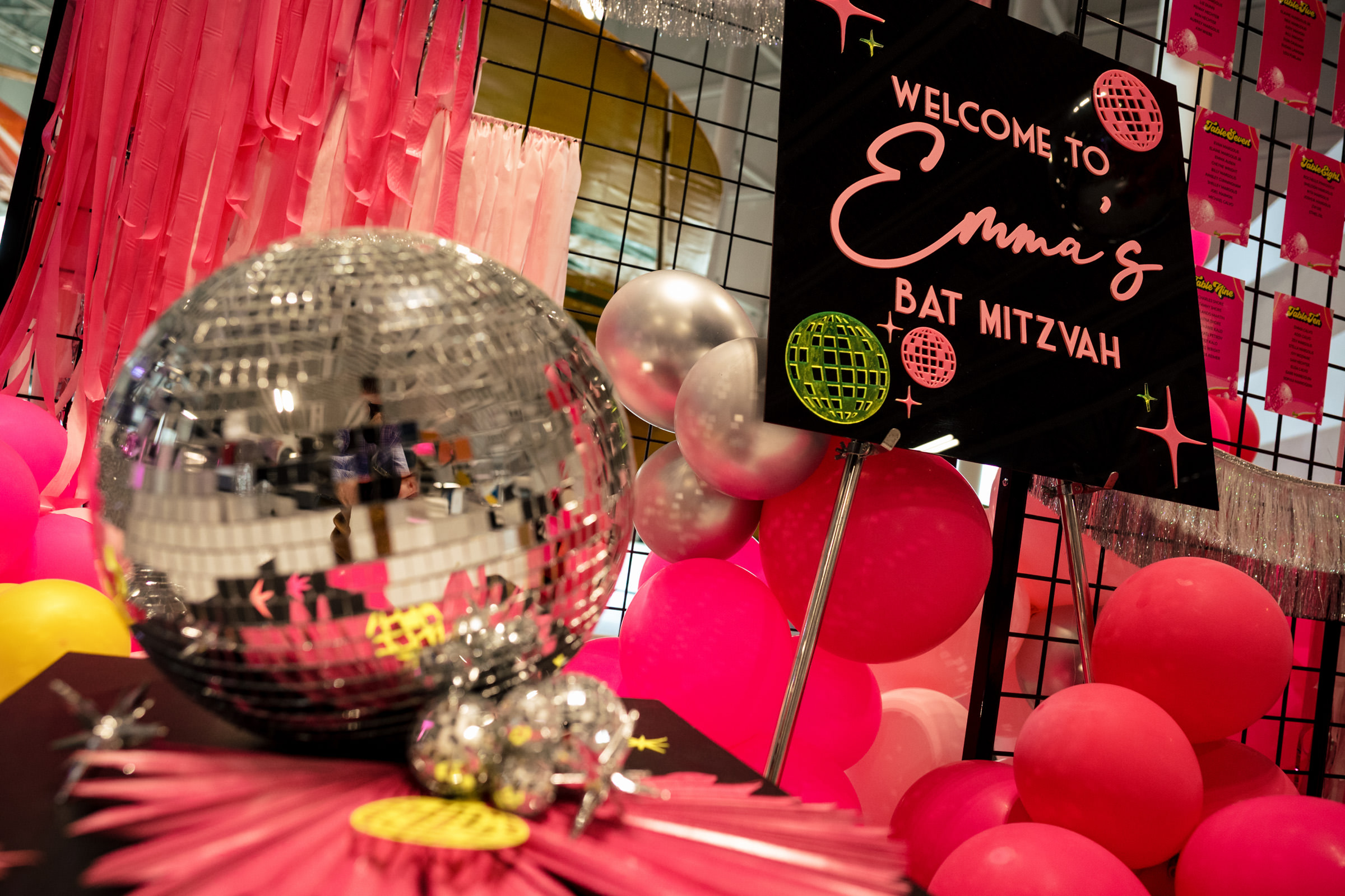 A Bat Mitzvah celebration is decorated with pink, yellow, and silver balloons, and metallic string curtains. A sign reads "Welcome to Emma's Bat Mitzvah." A disco ball centerpiece is prominently placed in front of the sign.