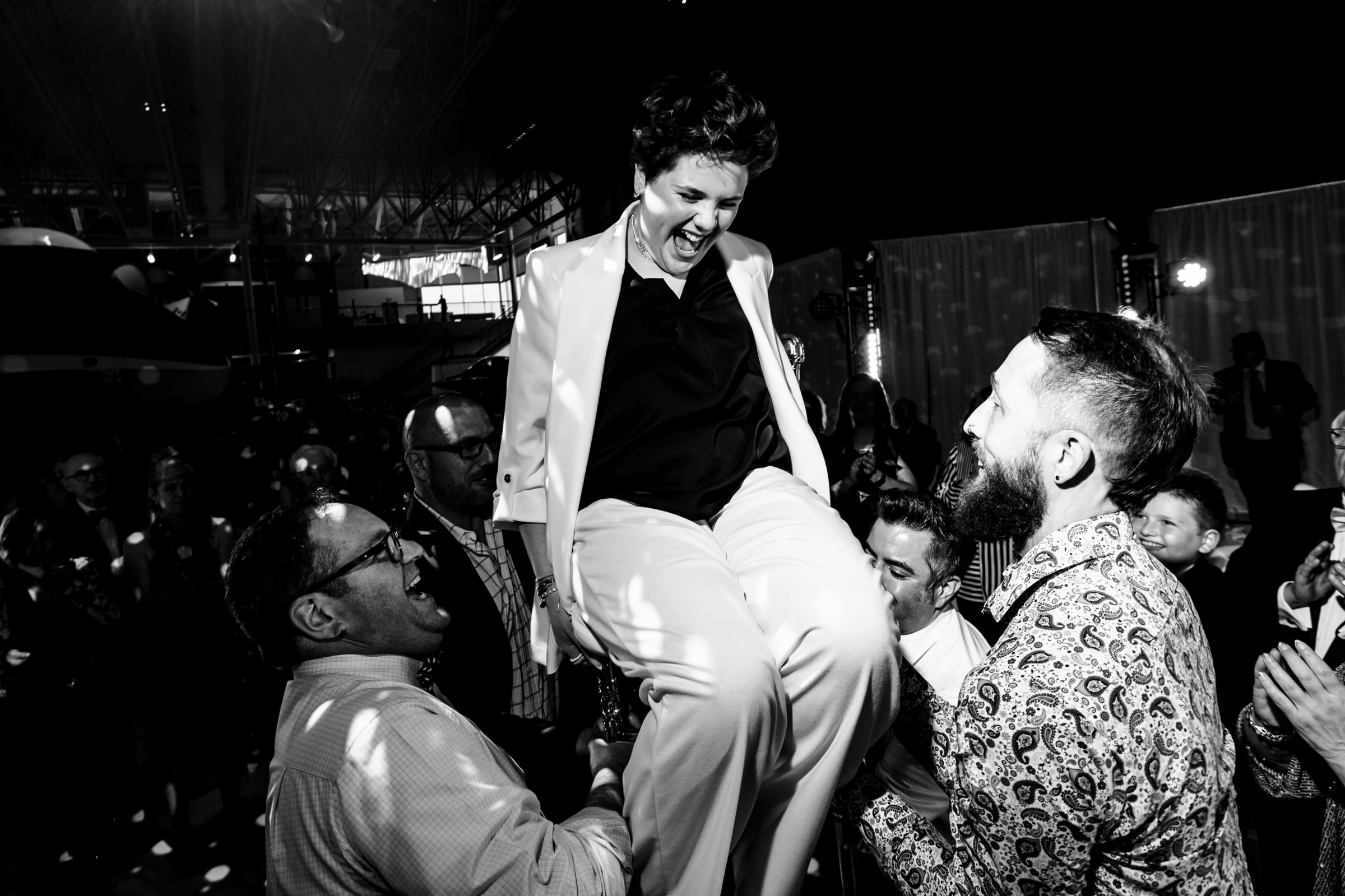 A black-and-white photo shows a joyful person dressed in a suit being lifted into the air by several others at a lively event. The group of people around them are smiling and appear to be celebrating. The background is dark, with lights and some visible structures.