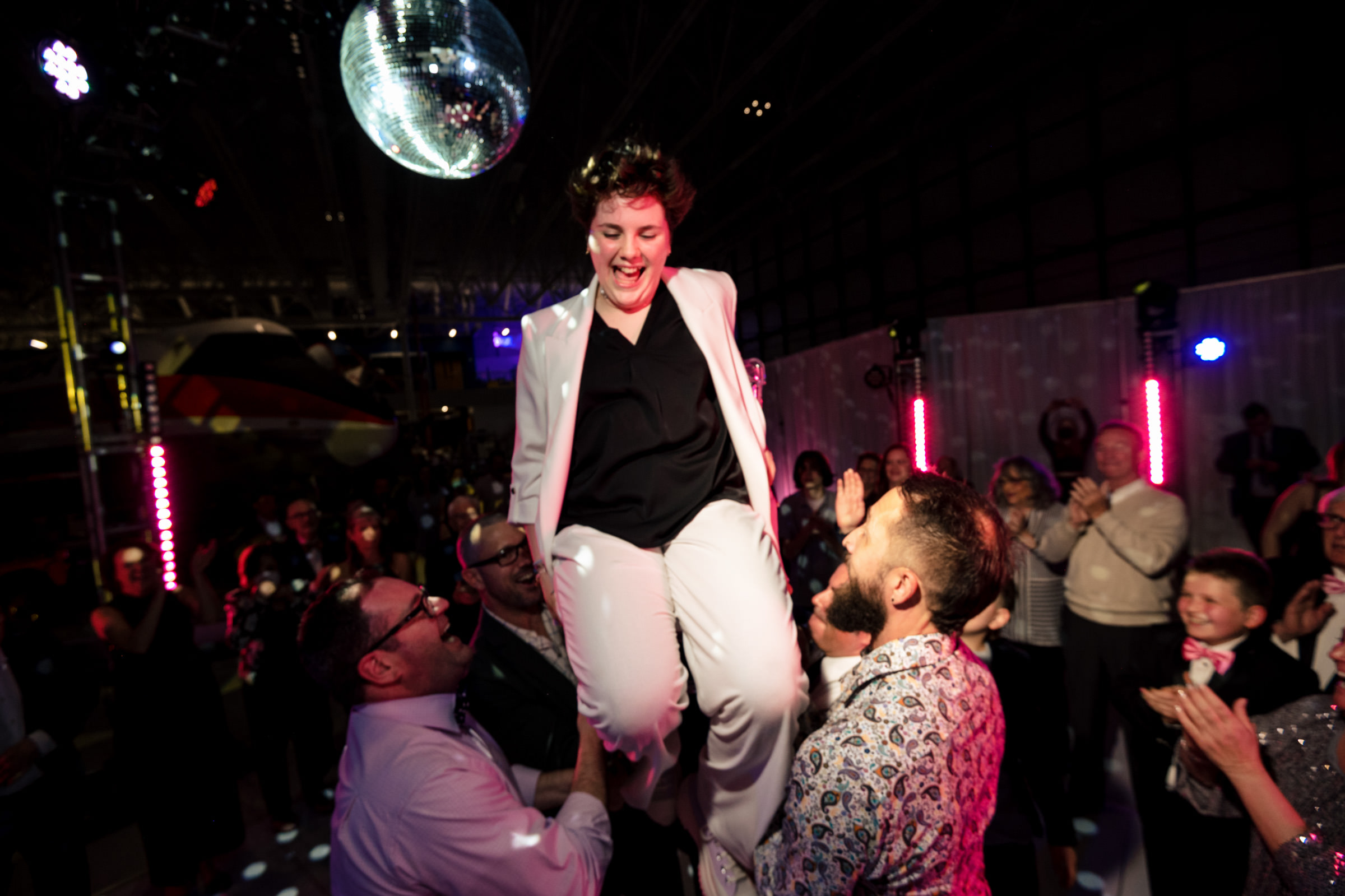 A person in a white suit is joyfully lifted into the air by two other individuals during a celebratory event. The scene is energetic, with a disco ball overhead, colorful lighting, and a crowd of people clapping and smiling in the background.