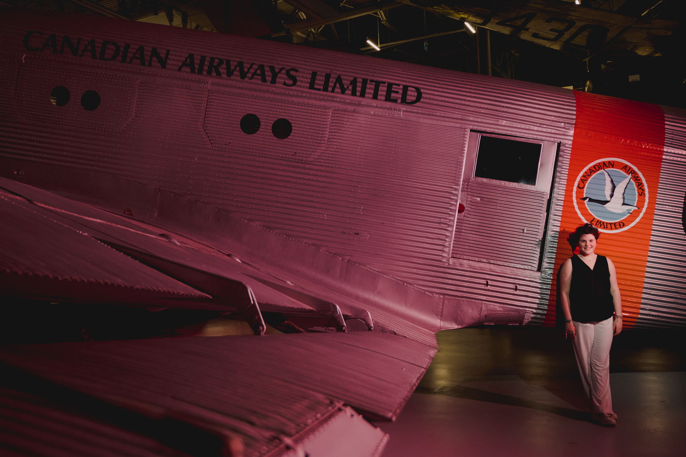 A person wearing a black jacket and white trousers is standing beside a large vintage airplane with "Canadian Airways Limited" written on the side. The aircraft is illuminated in red lighting, and a circular emblem is visible on the tail. The scene appears to be indoors.