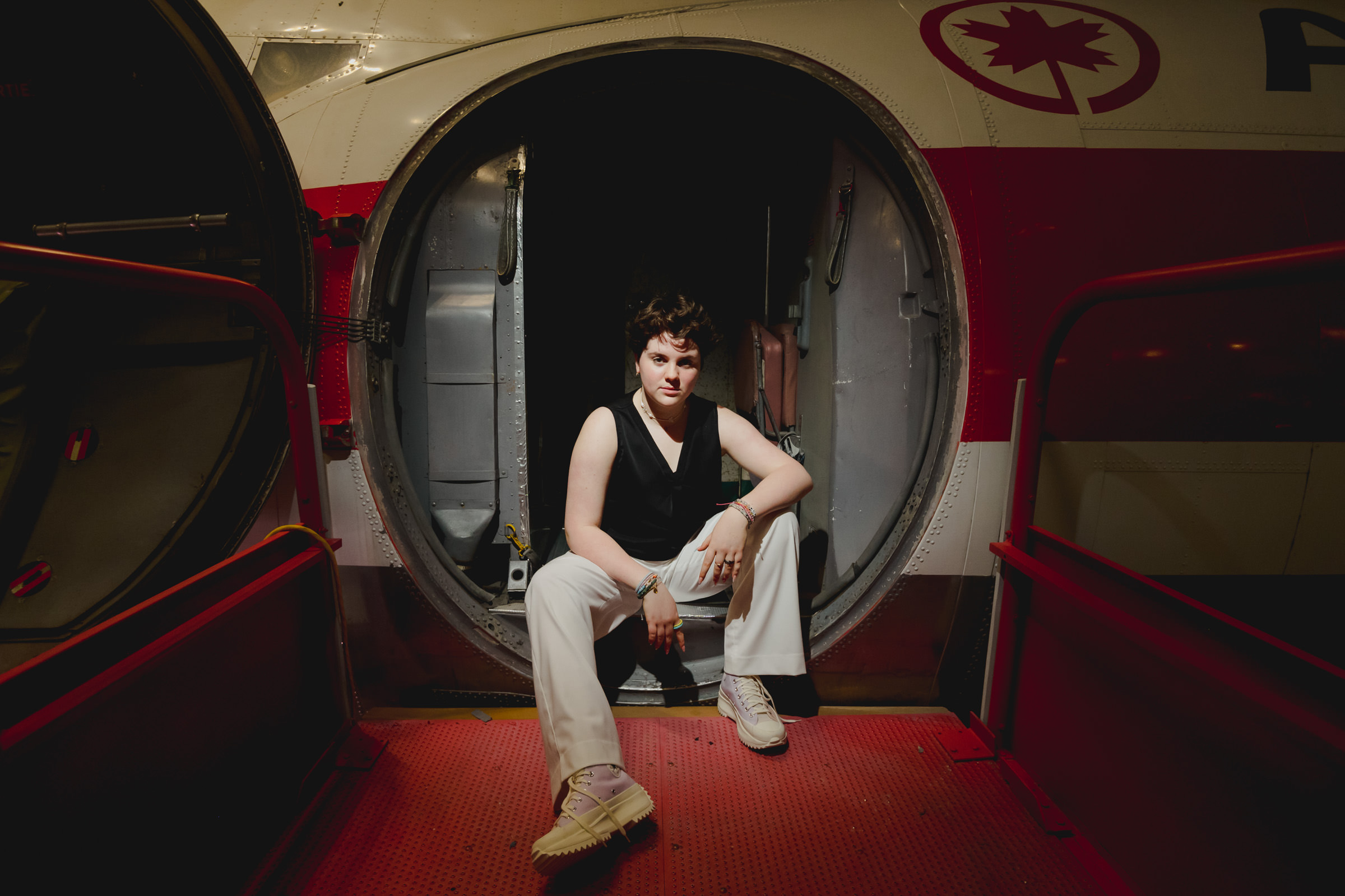 A person with short hair, wearing a sleeveless black top and white pants, sits confidently at the entrance of an aircraft with the door open. The interior and exterior of the aircraft are partially visible, and the floor around them is red.