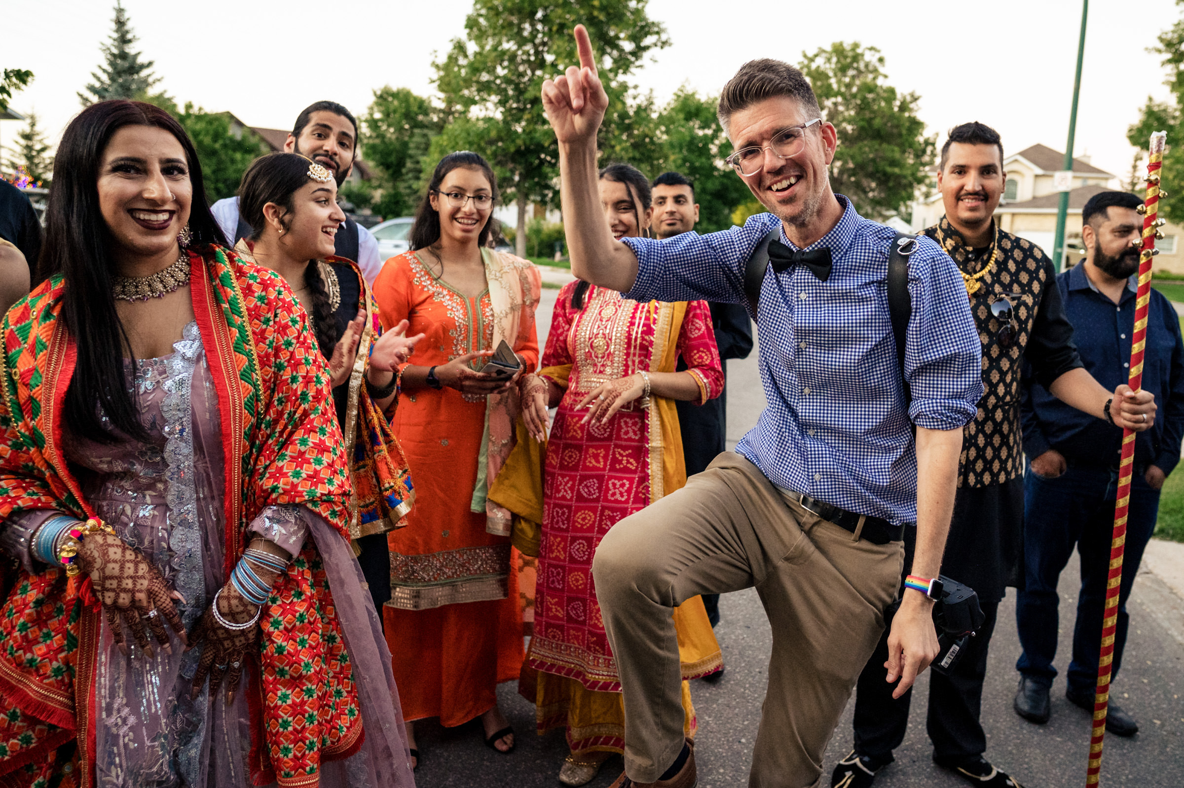 A group of people dressed in colorful traditional attire is smiling and celebrating outdoors. In the foreground, a man wearing glasses, a blue shirt, and a bow tie is joyfully raising his hand and lifting one leg. Trees and houses are visible in the background.