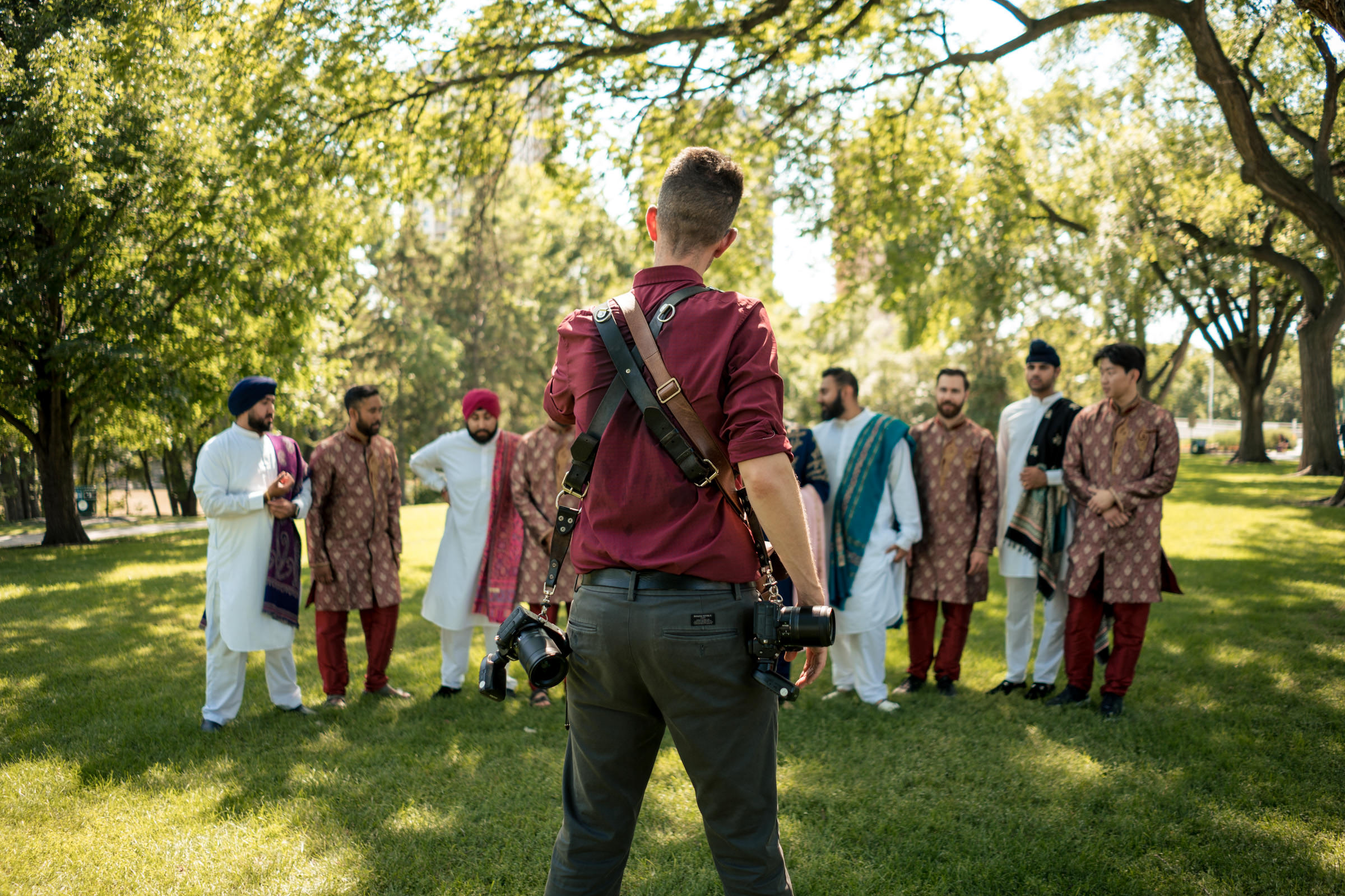 A photographer, holding cameras, stands in the foreground with his back to the camera. He captures a group of seven men dressed in colorful traditional attire, standing together under the shade of large trees in a park. The weather is sunny and the mood appears festive.