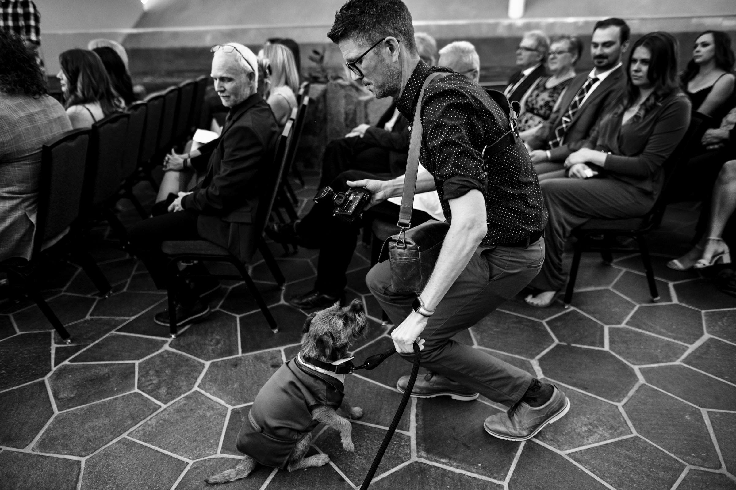 A man in glasses holding a camera steadies a dog on a leash, kneeling on the tile floor of a venue. The dog is wearing a vest. In the background, several people are seated, attentively watching something out of the frame. The scene is in black and white.