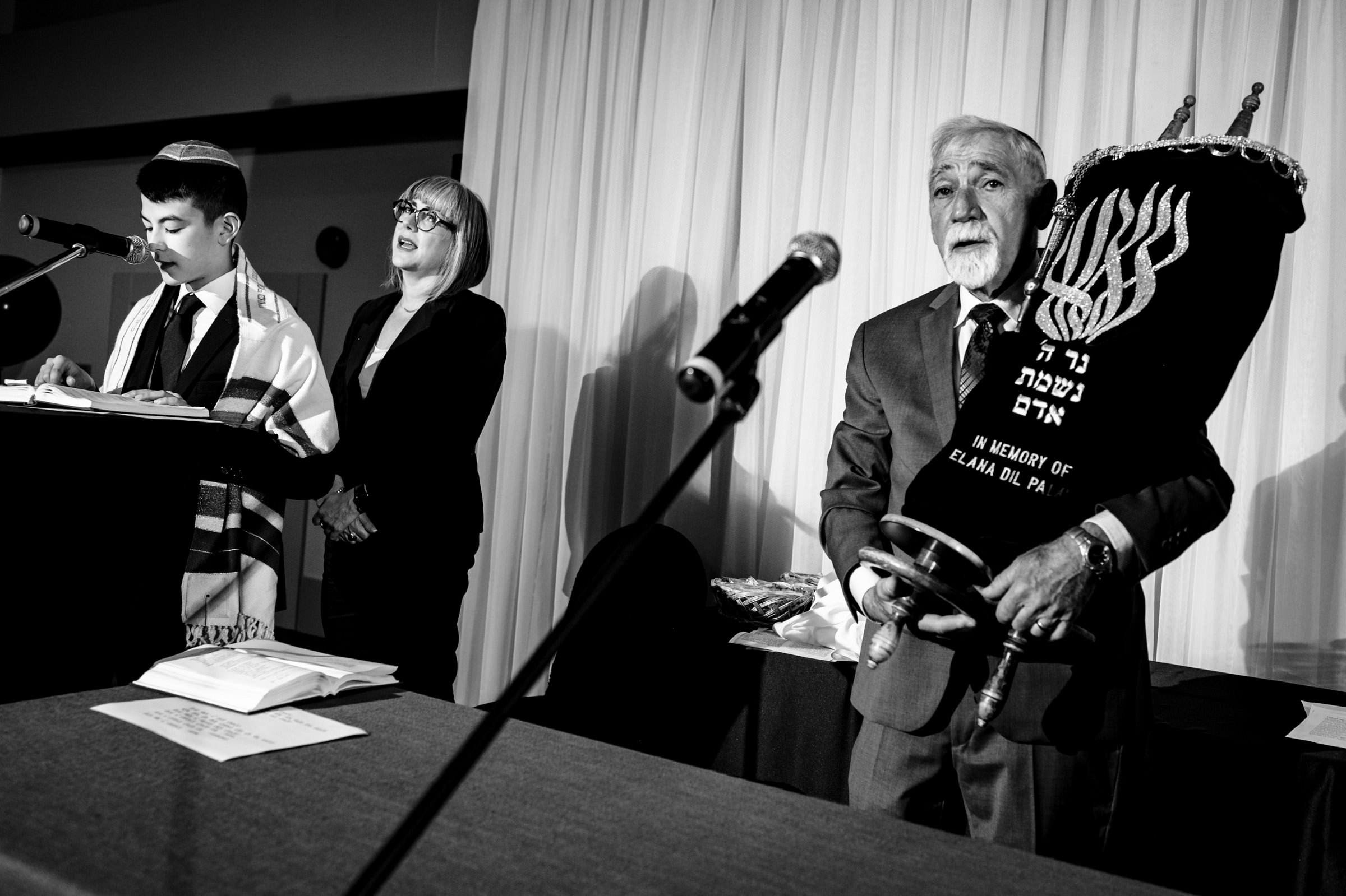 Black and white image of a religious ceremony. A boy stands at a podium with a prayer book, flanked by a woman singing and a man holding a Torah scroll. Two microphones are visible, and a Hebrew inscription is on the Torah cover.