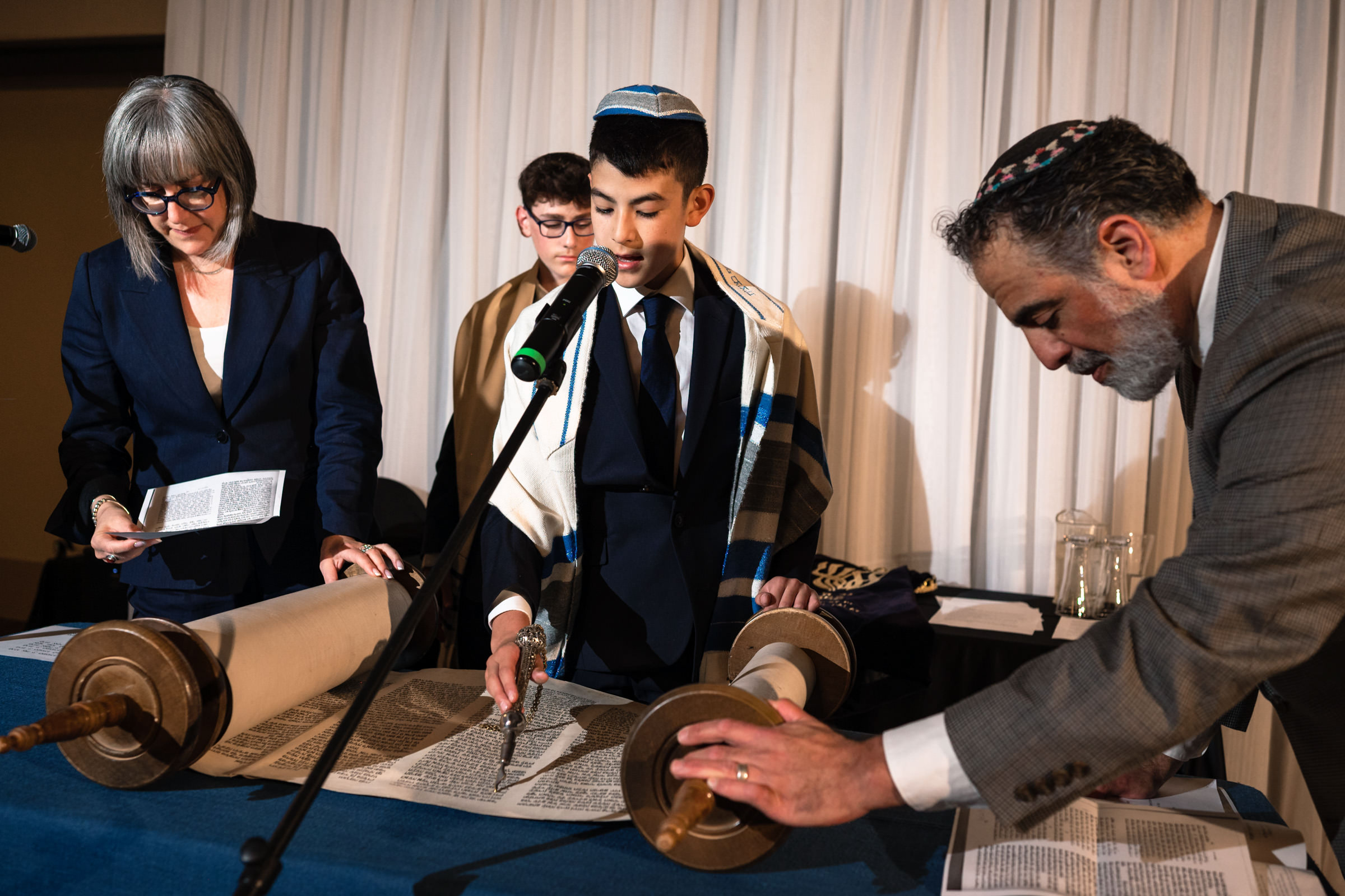A young boy, wearing a kippah and tallit, reads from the Torah during a Bar Mitzvah ceremony. An older woman to his left and an older man to his right assist, while another boy stands in the background. They are all dressed formally and focused on the Torah scroll.