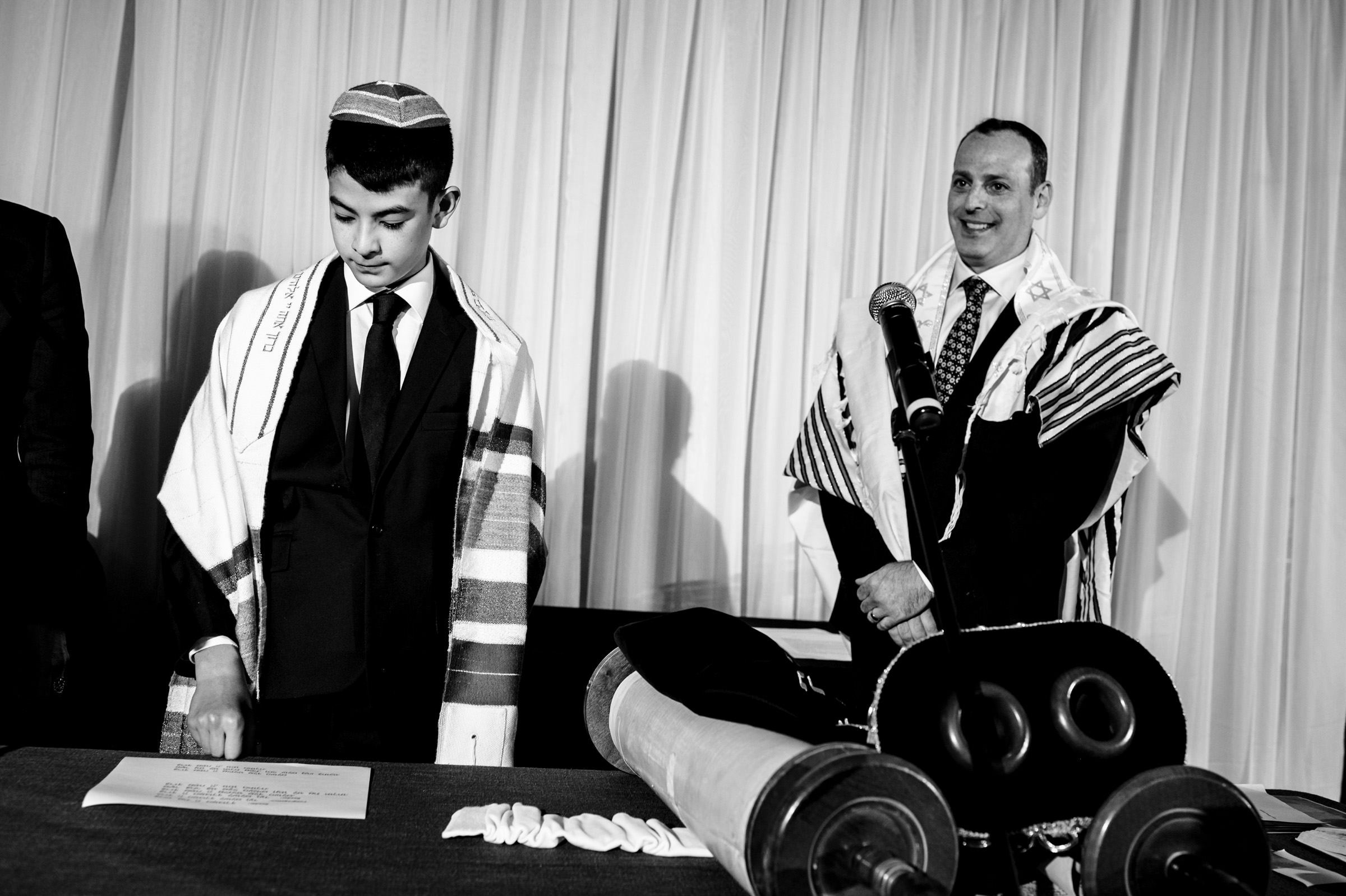 A young boy in a suit and a prayer shawl stands with a man wearing a similar shawl, both at a table with open religious scrolls and other items. The boy is looking down at a script, while the man smiles, holding a microphone.