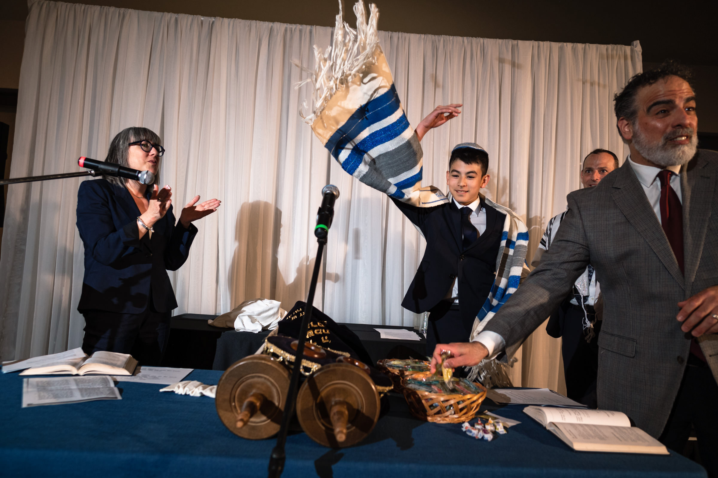 A young male is holding a tallit overhead during a religious ceremony. A woman with a microphone claps, while an older man in a suit is reaching for items on a table. A Torah scroll, prayer books, and a basket of items are laid out on the table.