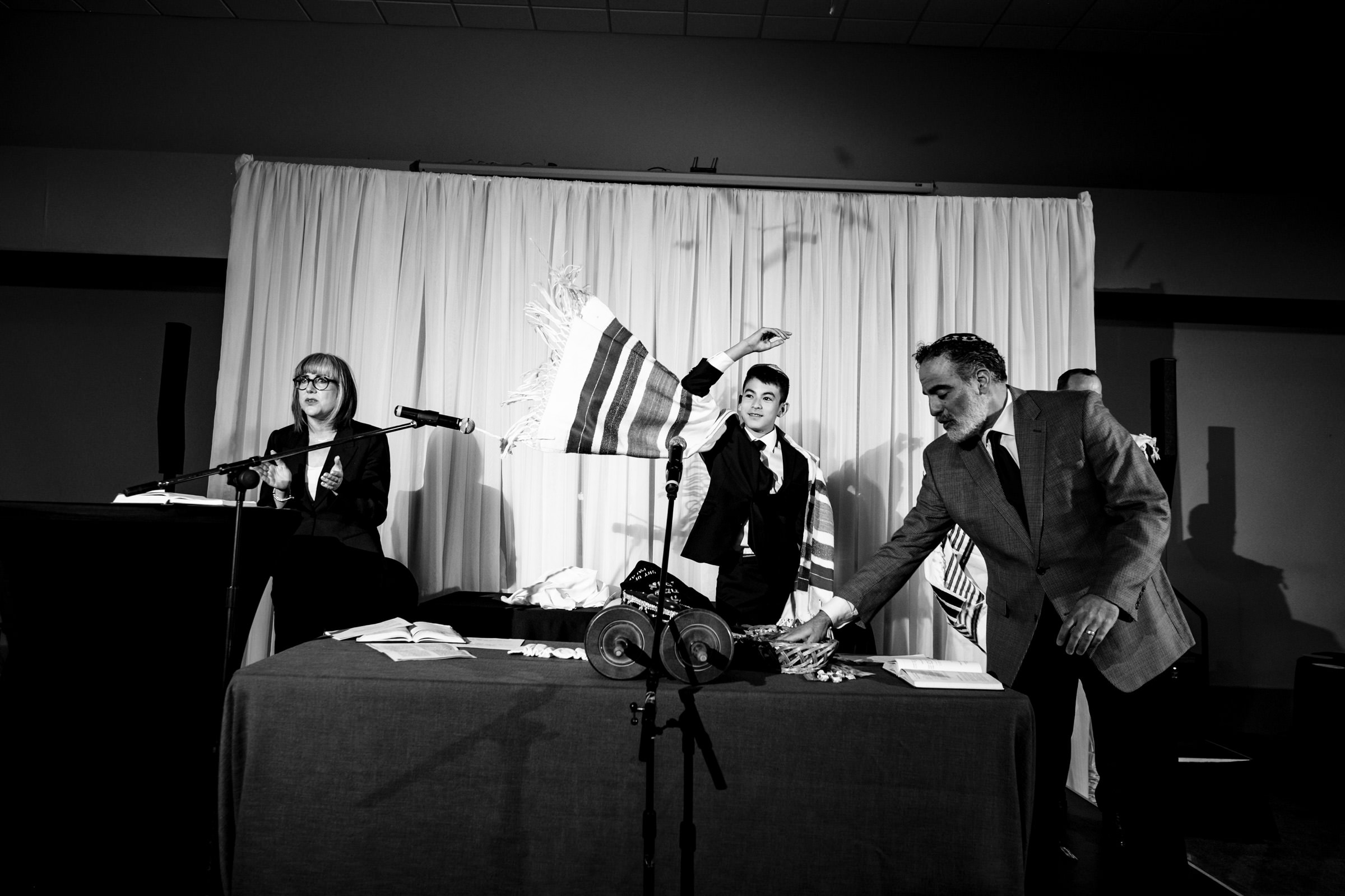 A black and white photo of three people in a room with a stage and curtain backdrop. One person on the left is speaking at a podium, the person in the middle is raising a fringed fabric, and the person on the right is reaching for an item on a table covered with a tablecloth.