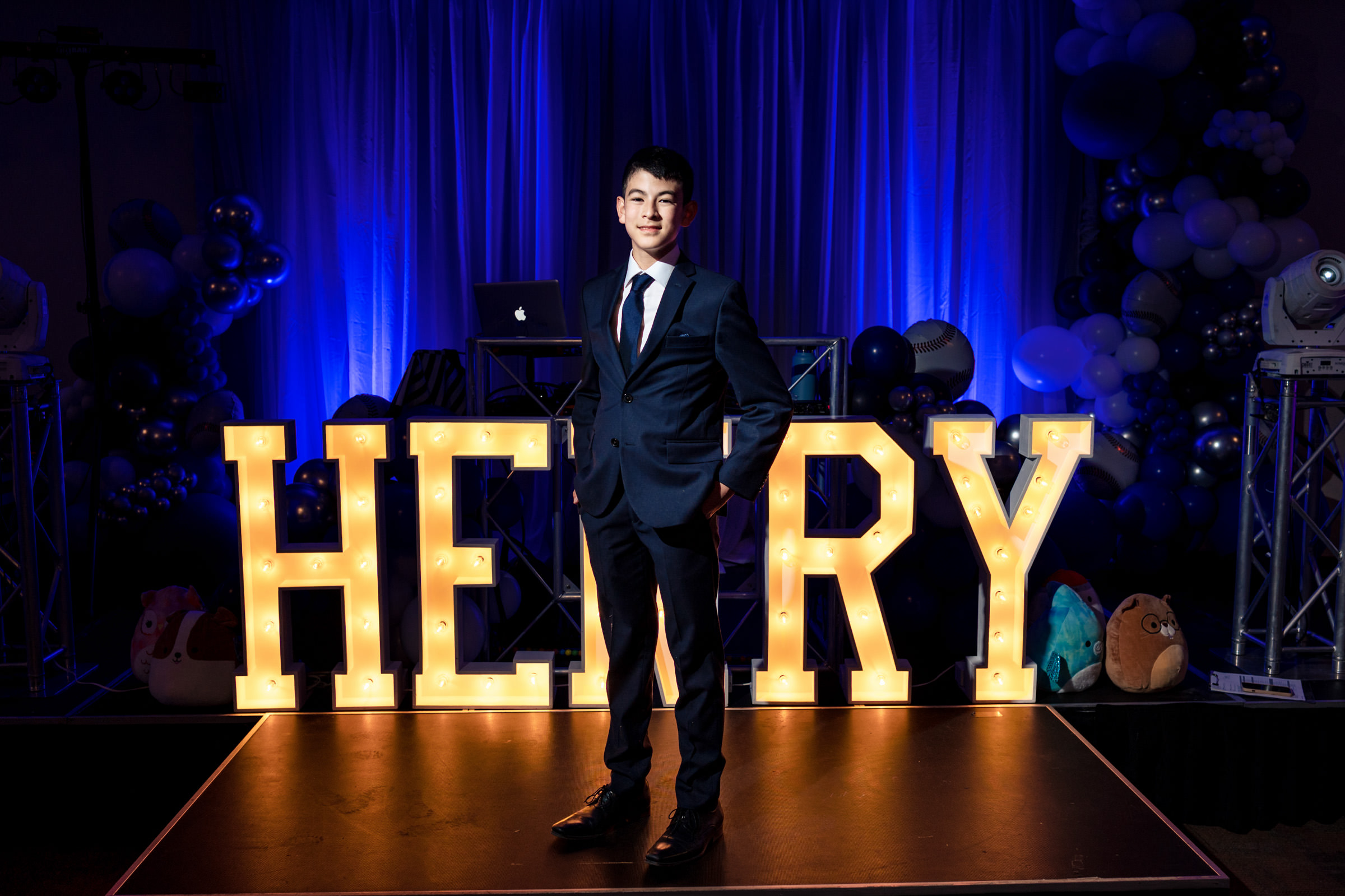 A young man in a navy suit and tie stands confidently on a stage with illuminated letters spelling "HENRY" behind him. The background is decorated with blue curtains, balloons, and various lights, creating a celebratory atmosphere.