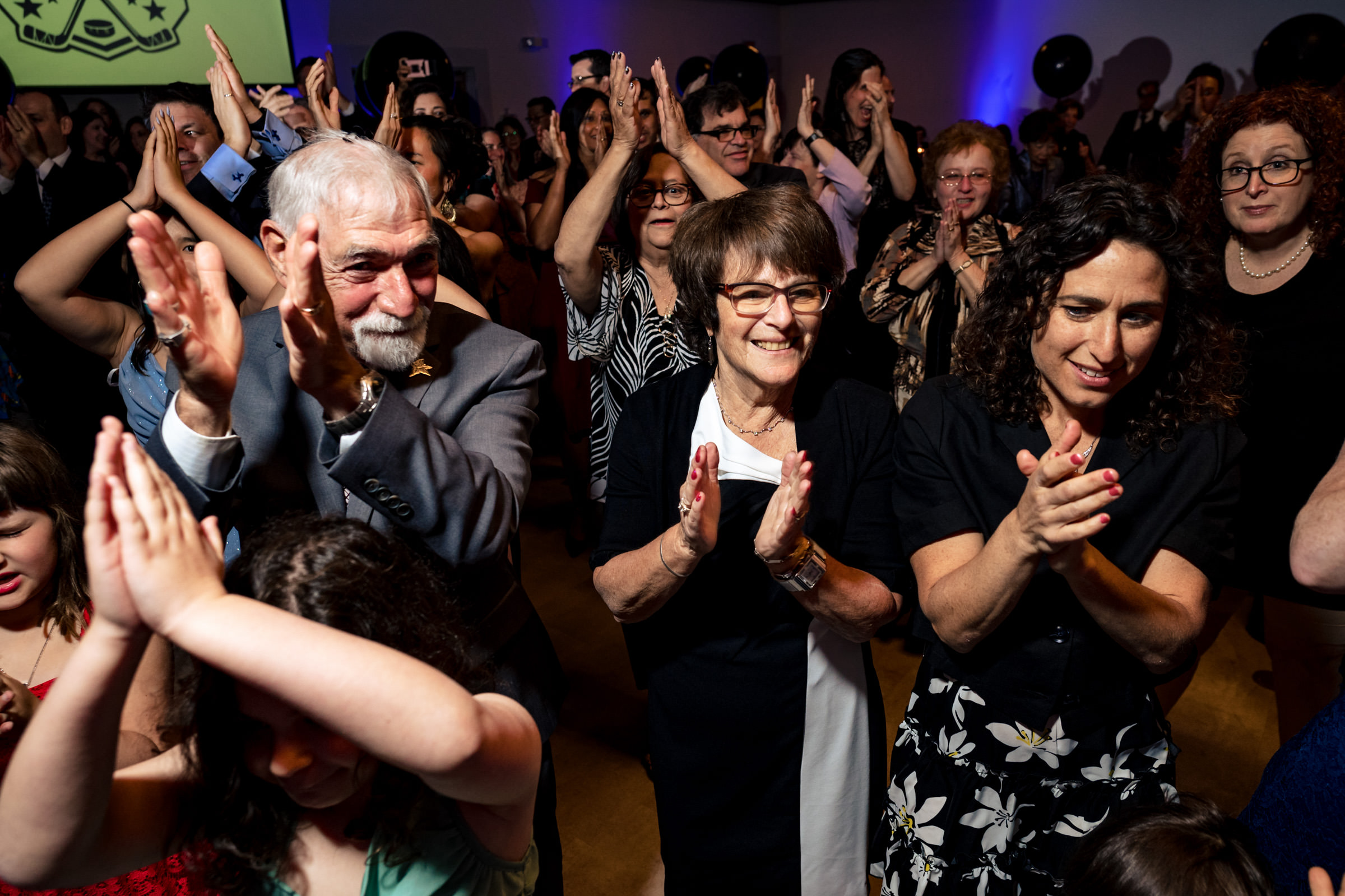 A diverse group of people of varying ages are happily clapping and dancing at a festive indoor event. The room is decorated with colorful lighting, and everyone appears to be enjoying themselves.