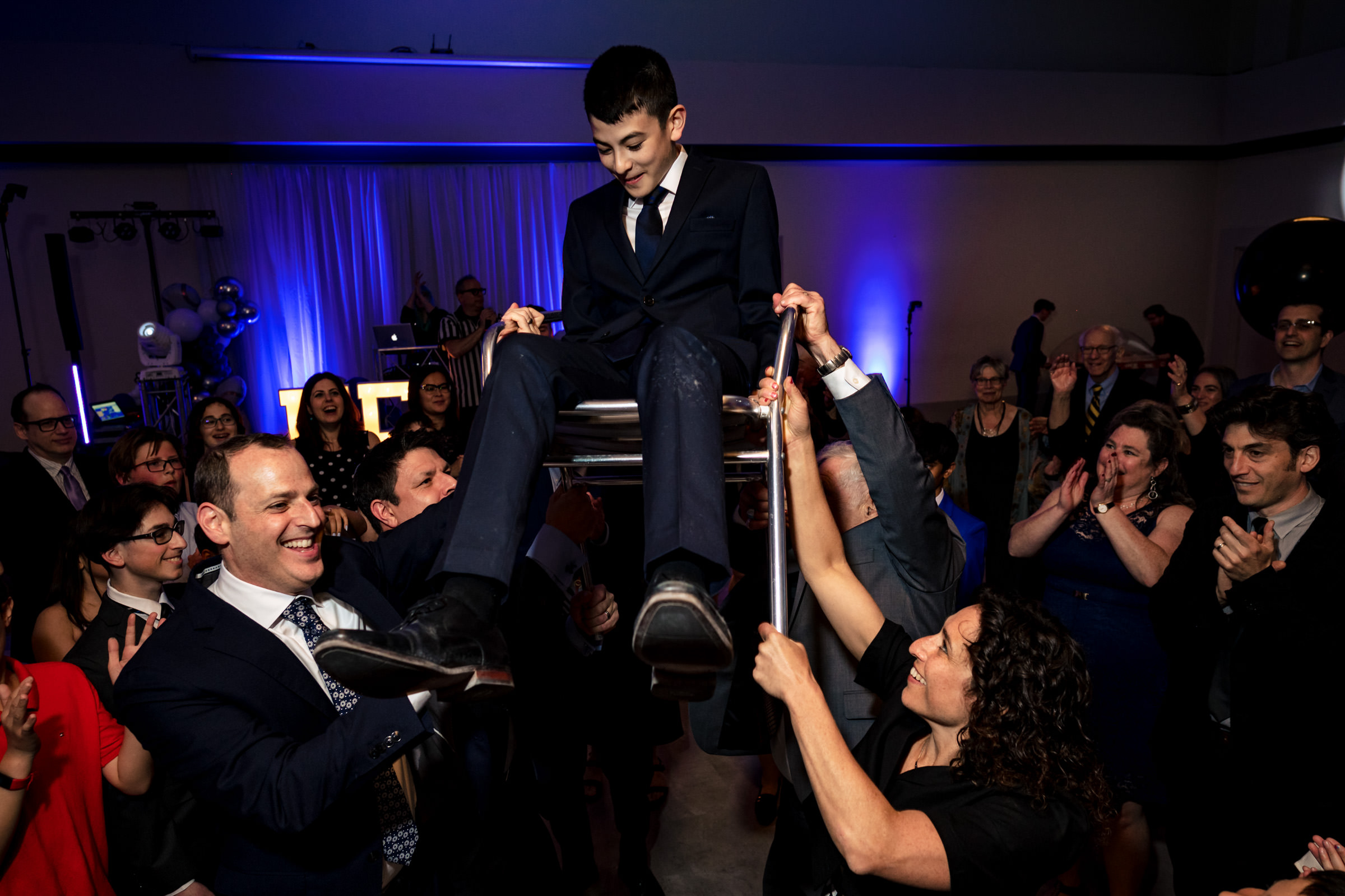 A boy in a suit is lifted on a chair by several people during a celebration. The crowd around him cheers and claps, with colorful lighting in the background and a DJ in the distance. Everyone appears joyful and engaged in the festive atmosphere.