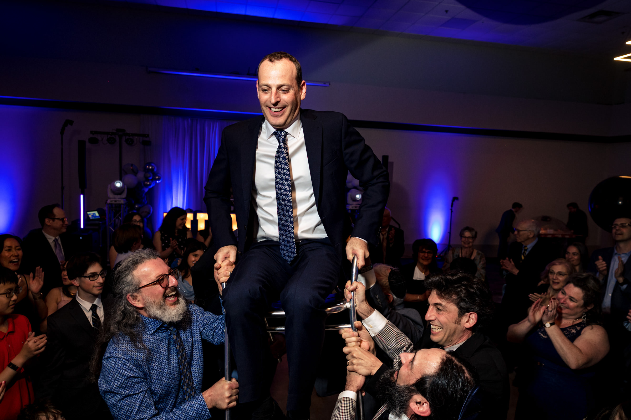 A man in a suit is lifted in a chair by three others during a celebration in a dimly lit room with blue and purple lighting. The room is filled with people clapping and smiling.